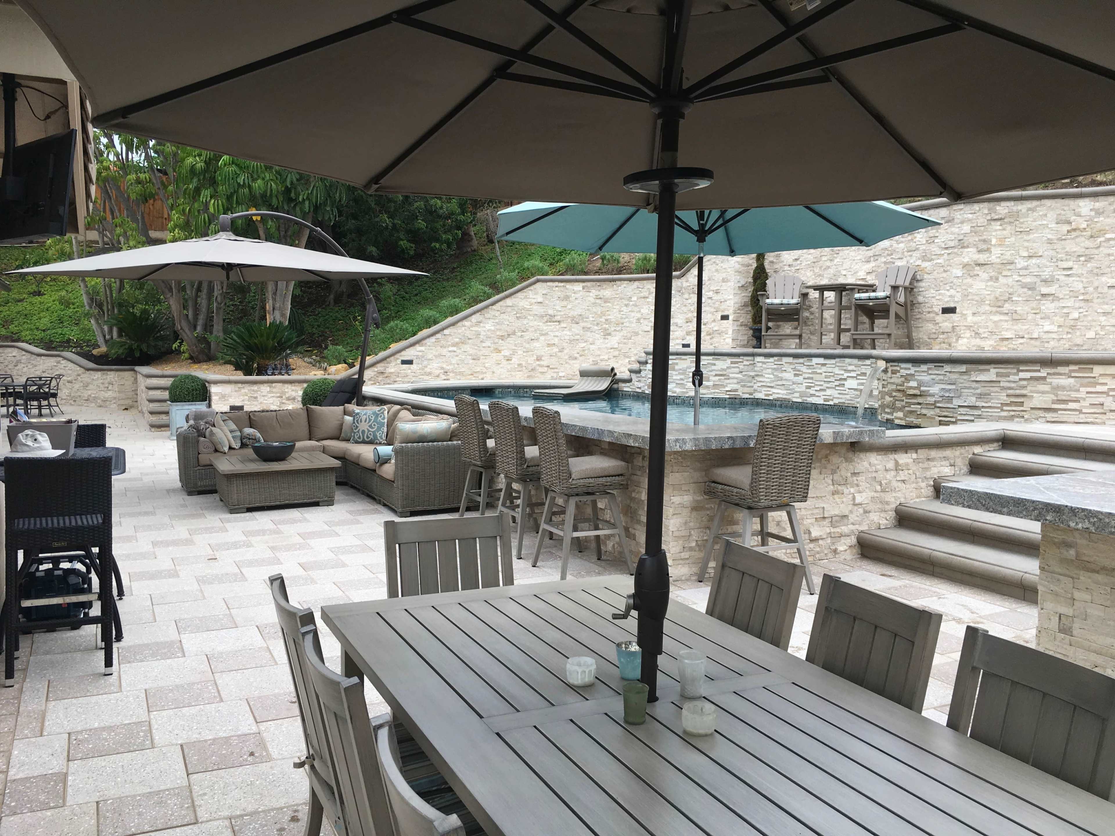 The image shows a patio area featuring outdoor furniture, an umbrella, a swimming pool, and a stone wall landscape.