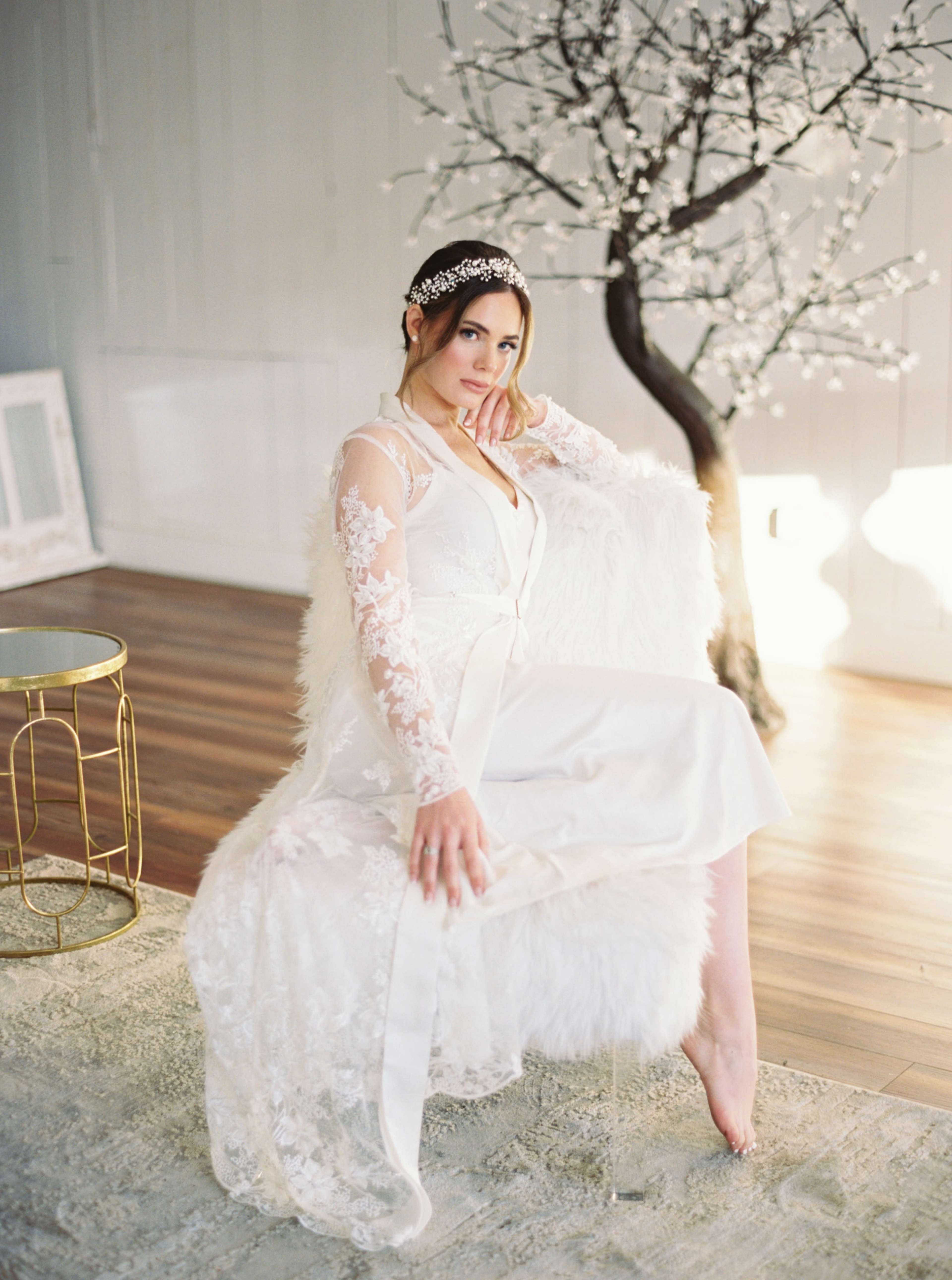 A woman in a white gown sits elegantly on a plush chair near a decorative tree with blossoms in a softly lit room.