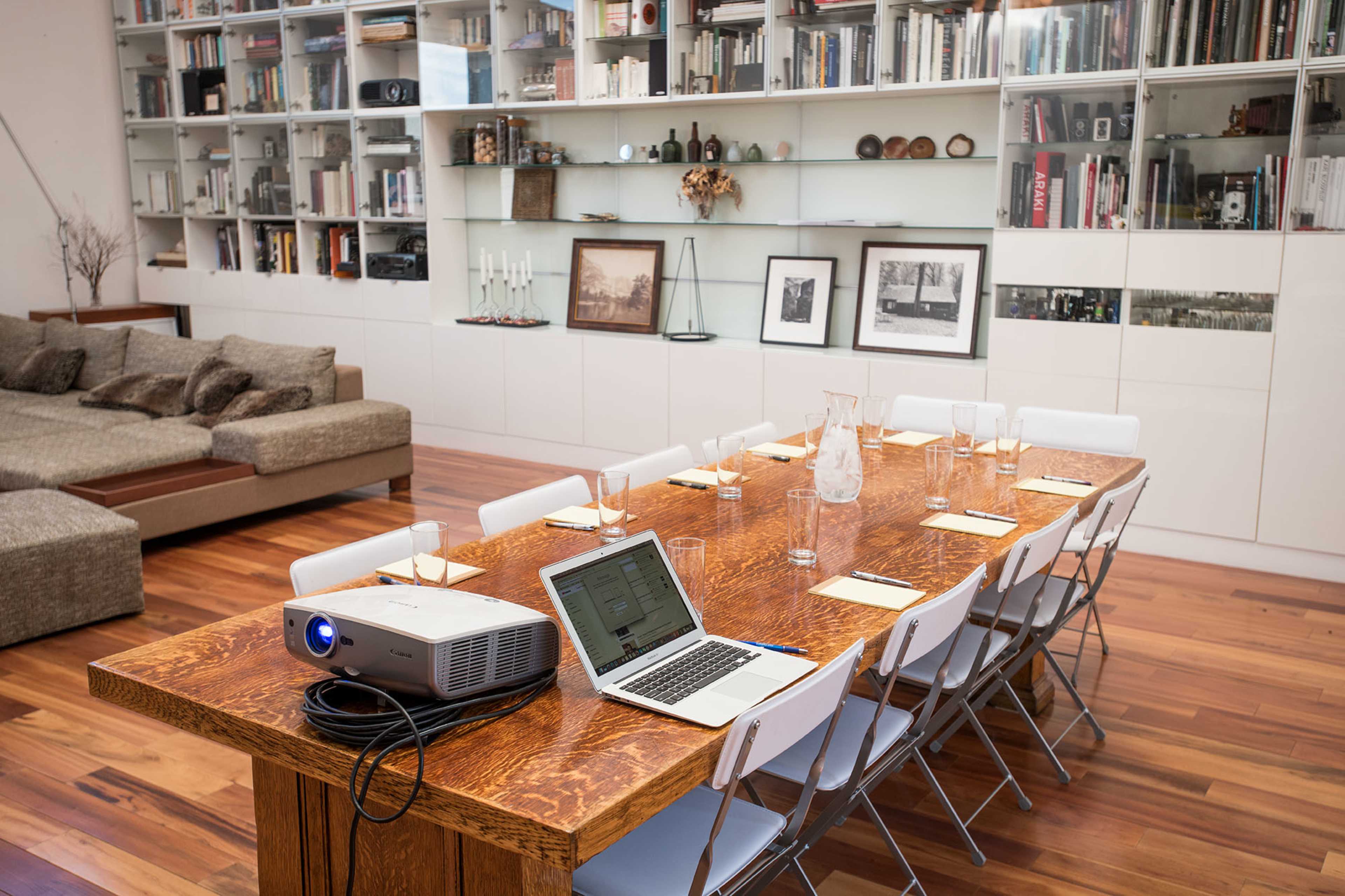 A long wooden table is set with glasses and notepads, accompanied by a laptop and projector in a spacious room featuring a large bookshelf.