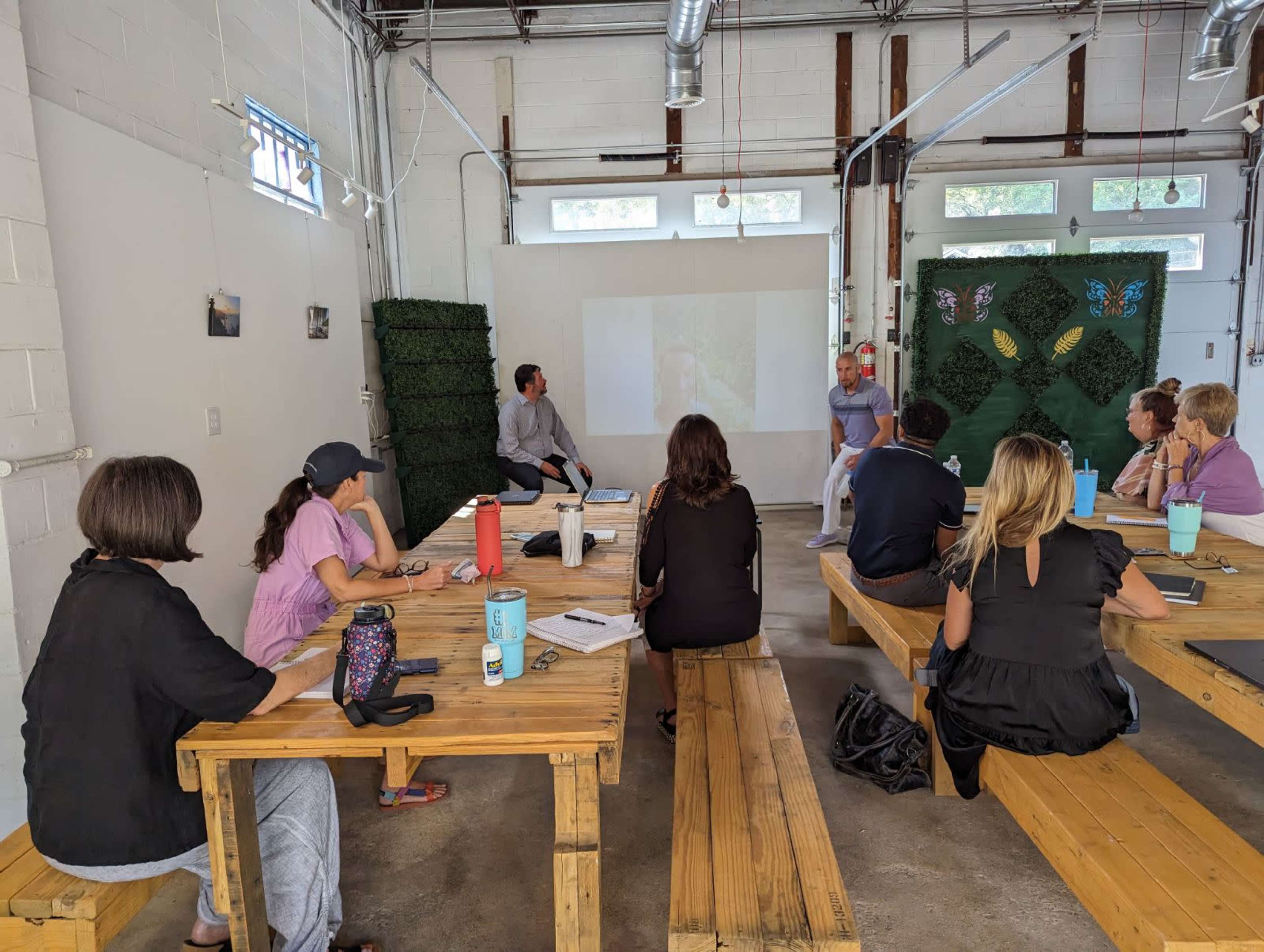 A group of people is seated at wooden tables in a bright room, listening to a speaker in front of a projected image.