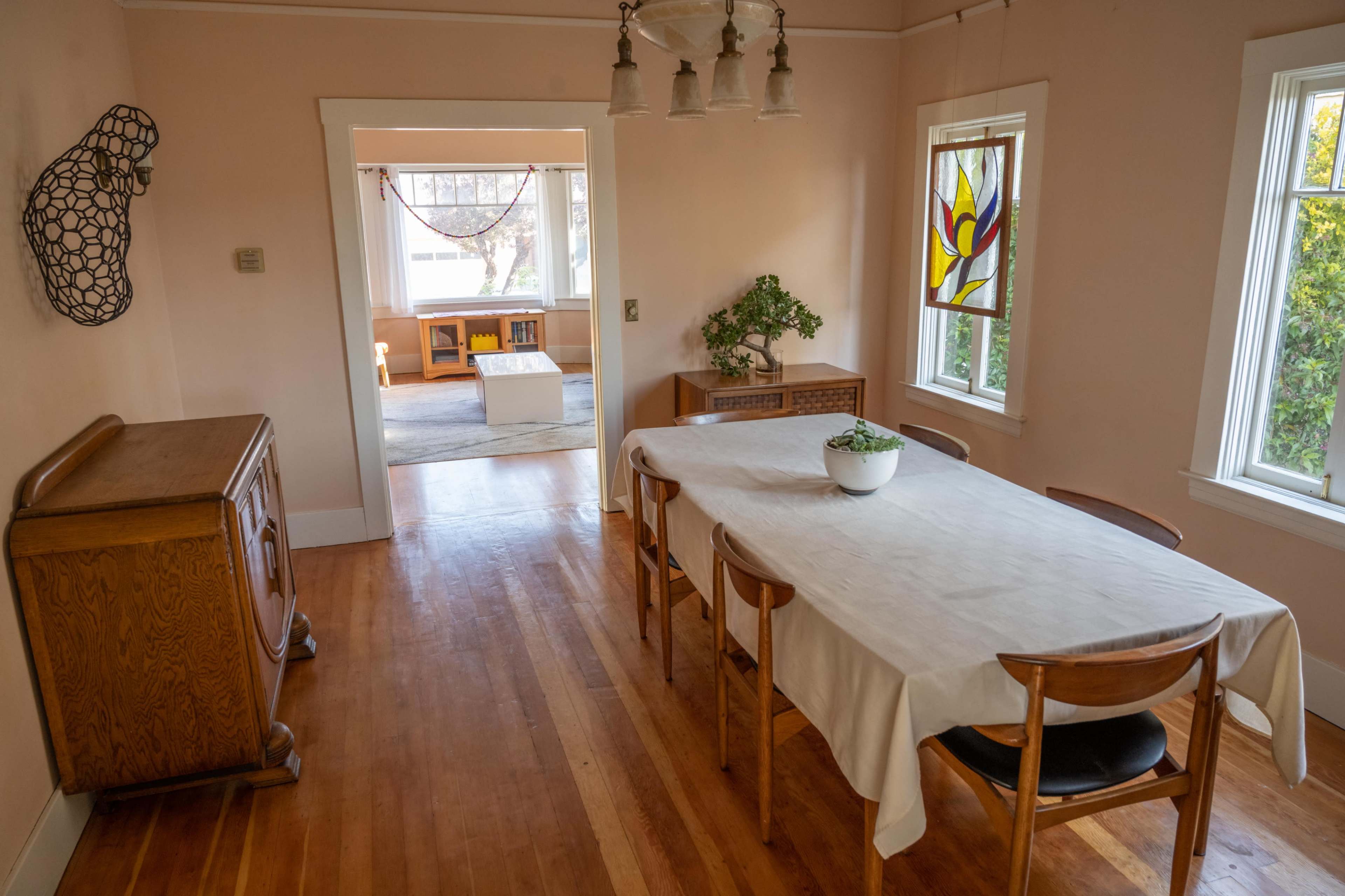 A dining room with a wooden table, chairs, and a sideboard, leading to an adjoining room with a light-colored floor and a decorative window.