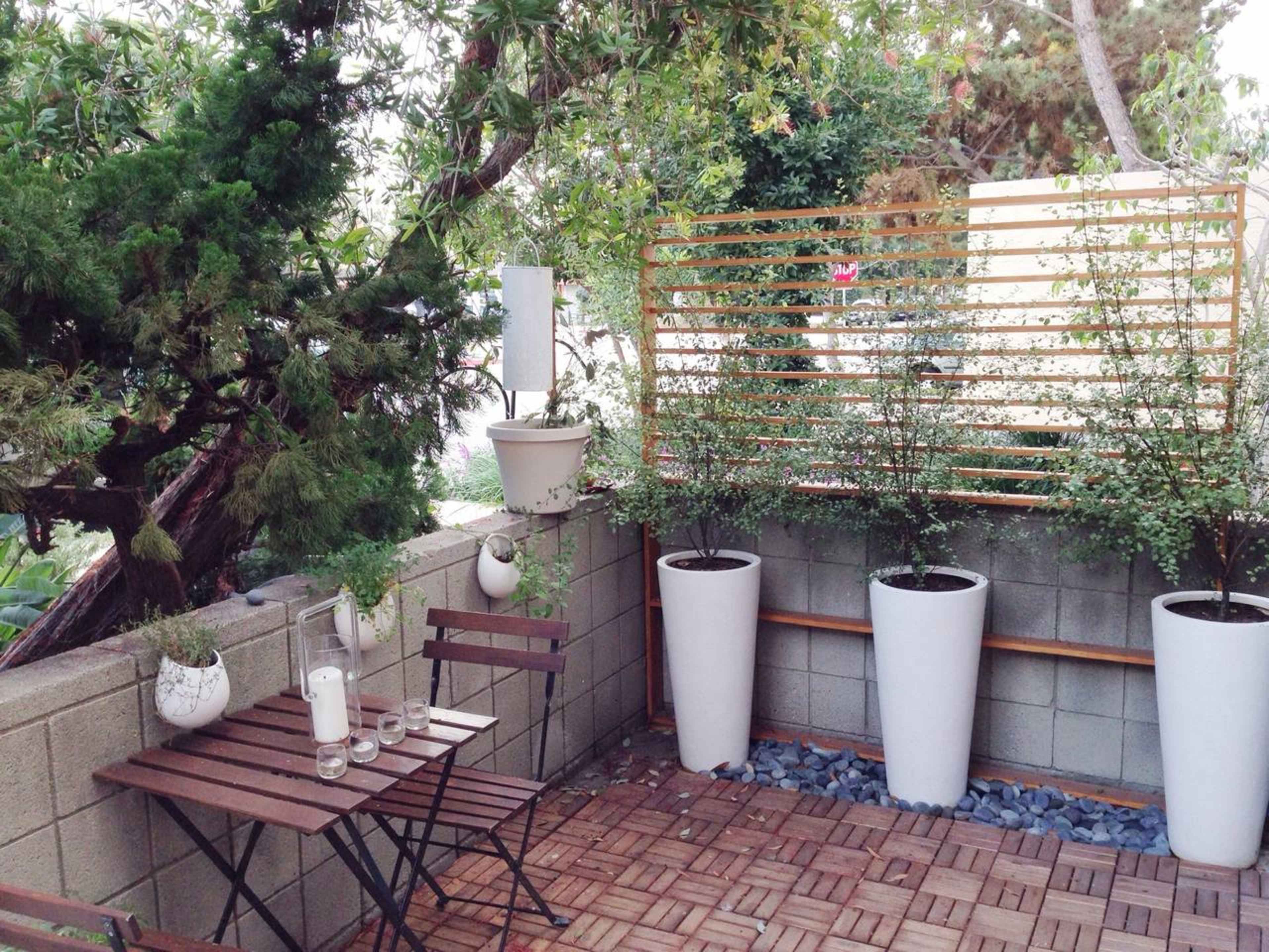 A small outdoor garden area featuring a wooden table and chairs, along with potted plants against a textured wall.