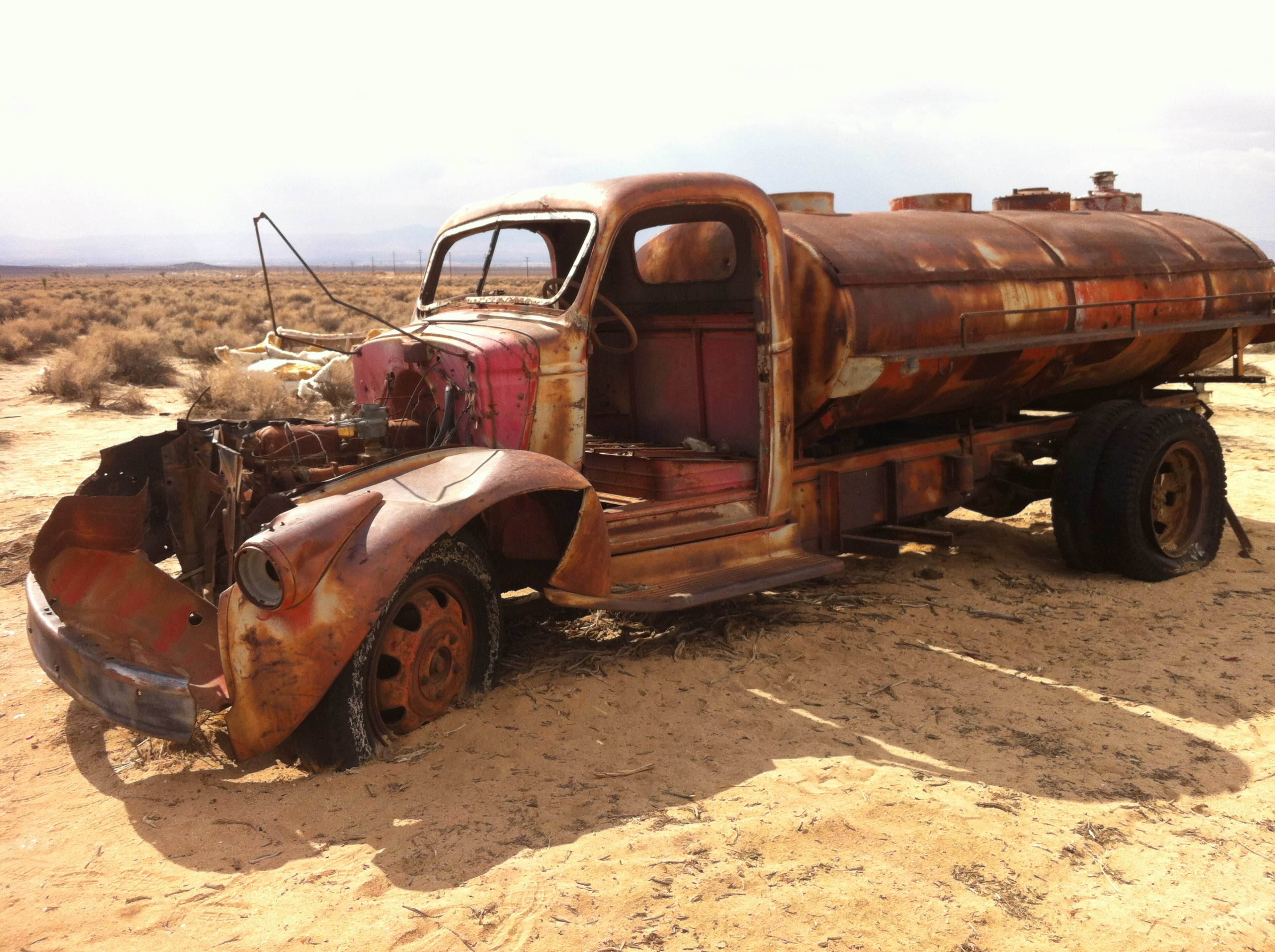 An old, rusted tanker truck is abandoned in a sandy, arid landscape with sparse vegetation.