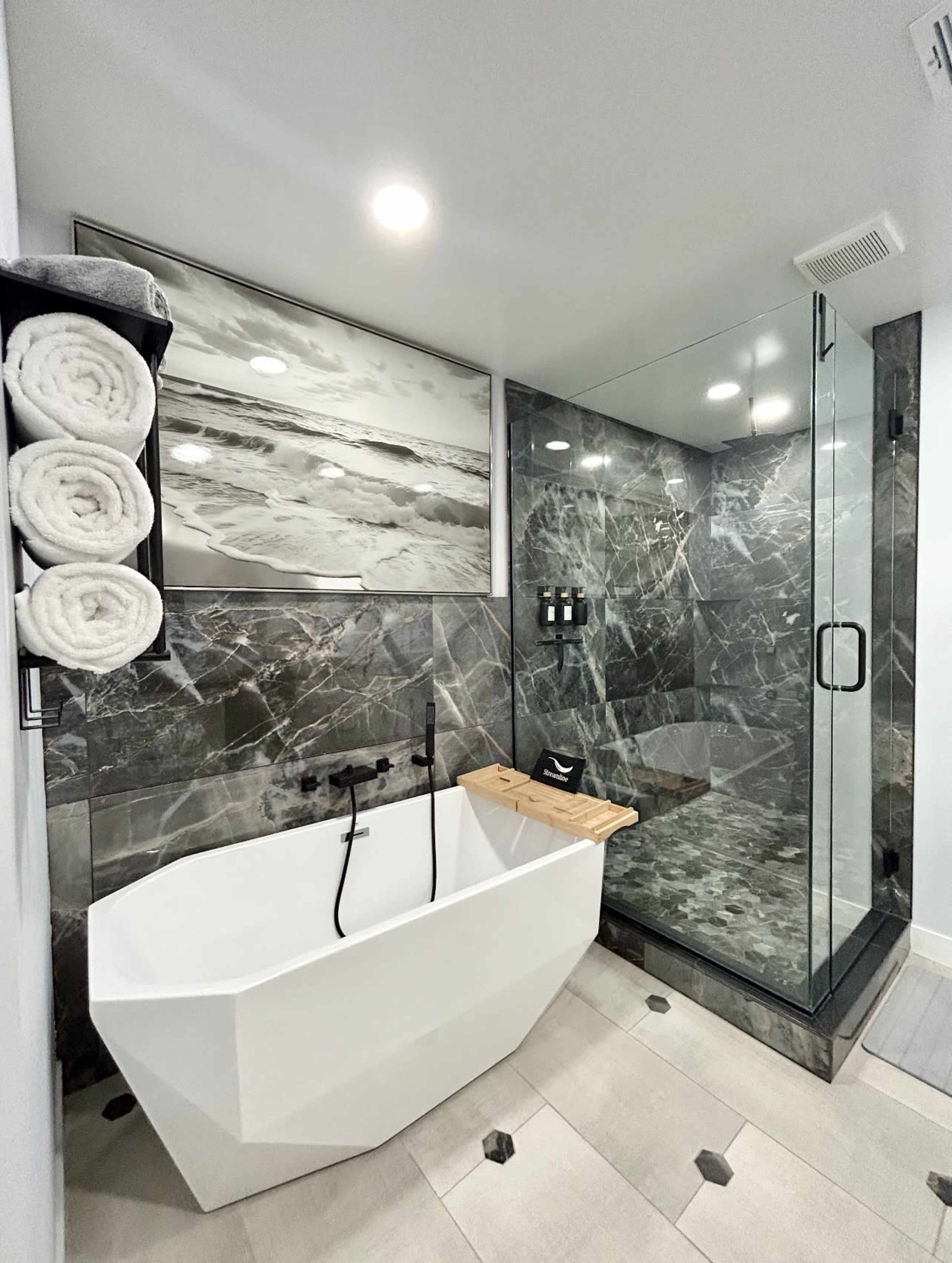 The image shows a modern bathroom featuring a freestanding white bathtub, a glass shower enclosure, and dark marble walls with a framed landscape photo above the tub.