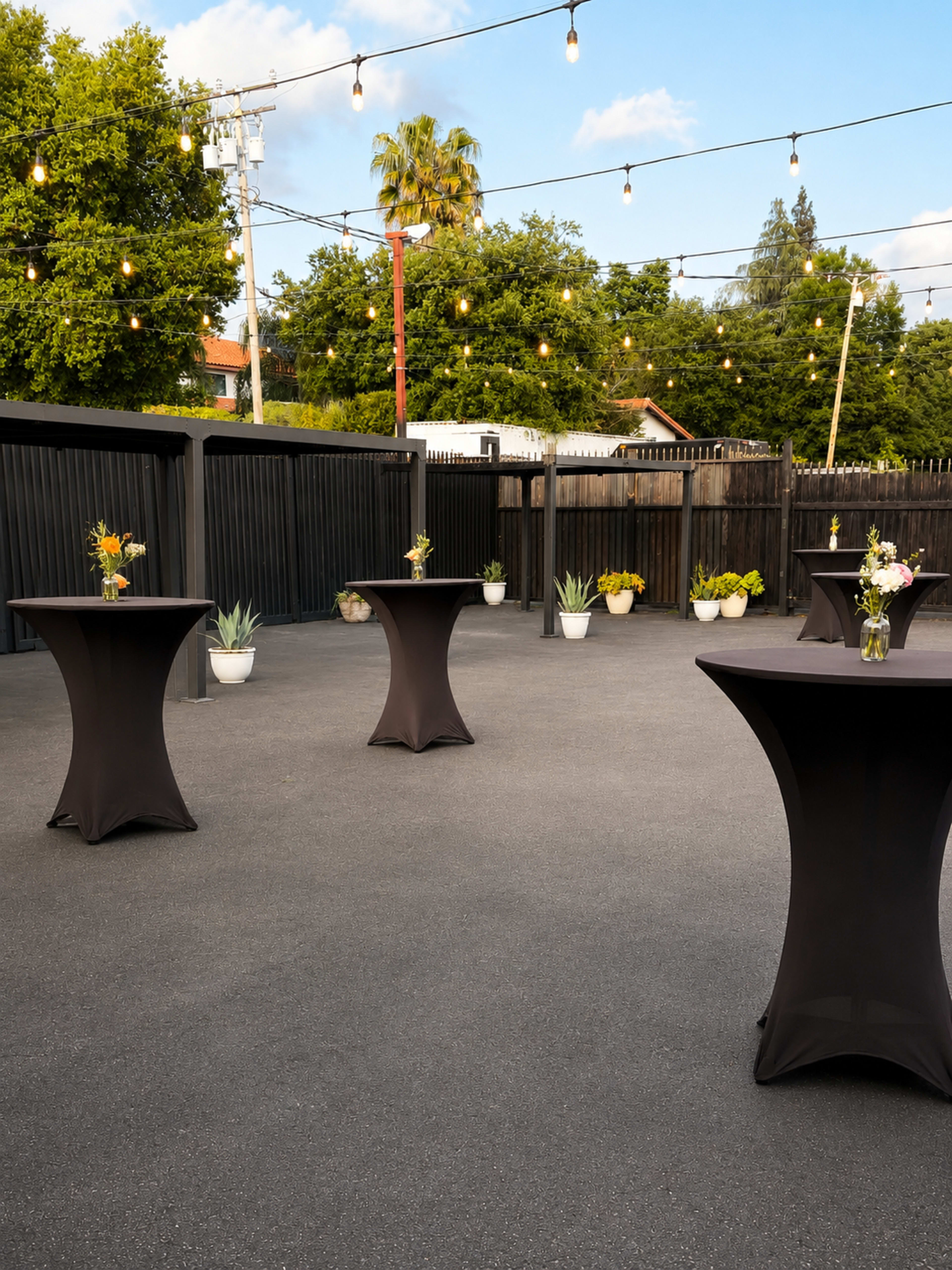 The image shows an outdoor event space with multiple round tables draped in black tablecloths, decorated with flowers, and string lights overhead.