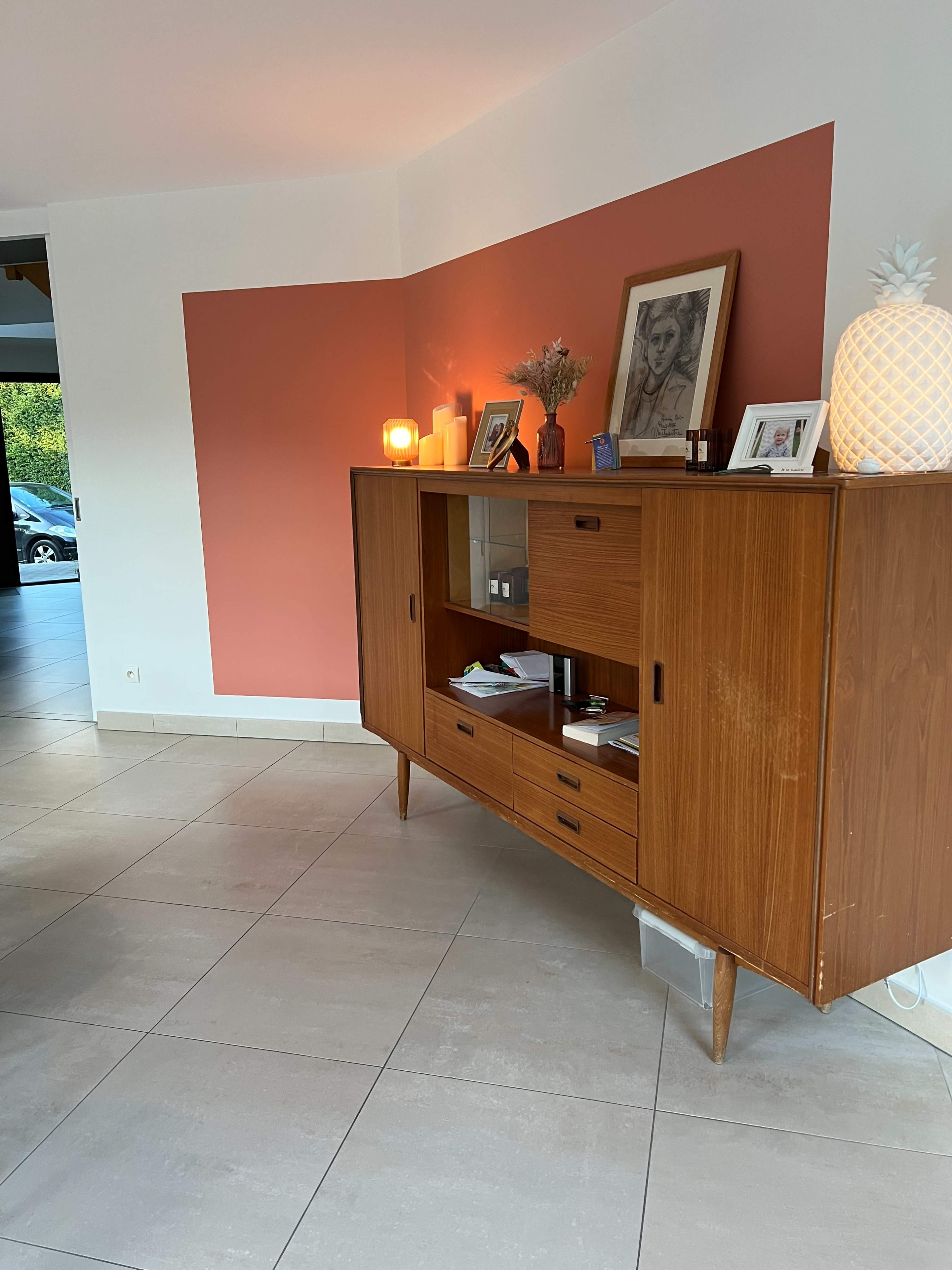 The image shows a corner of a modern living room with a wooden sideboard, decorative items, and a wall painted in coral against a neutral background.