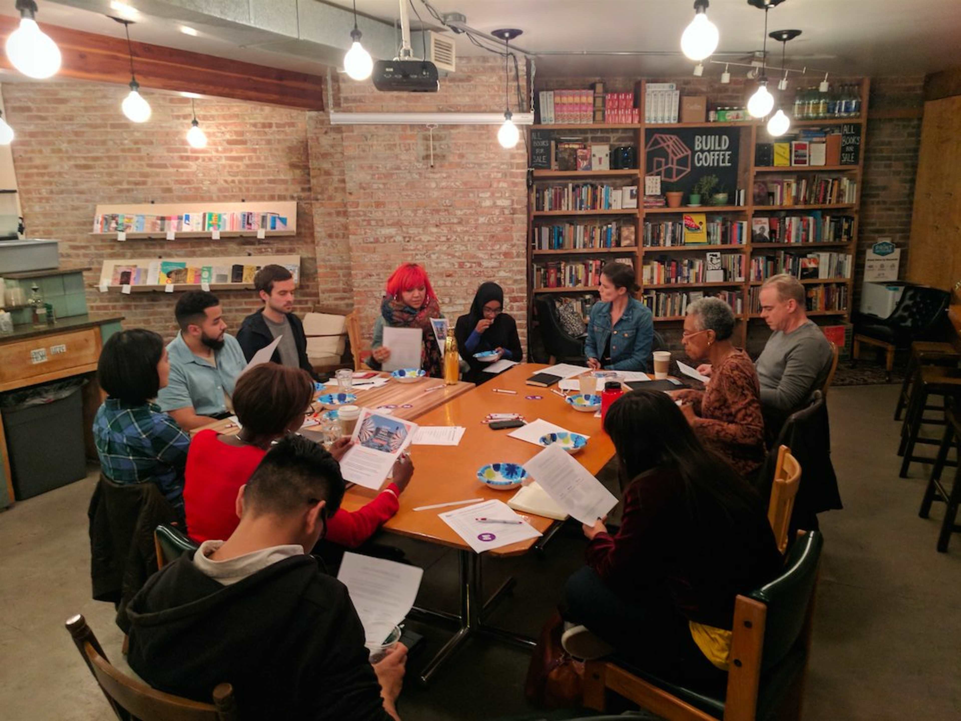 A group of diverse individuals is seated around a large table in a cozy, book-lined space, participating in a meeting or discussion.