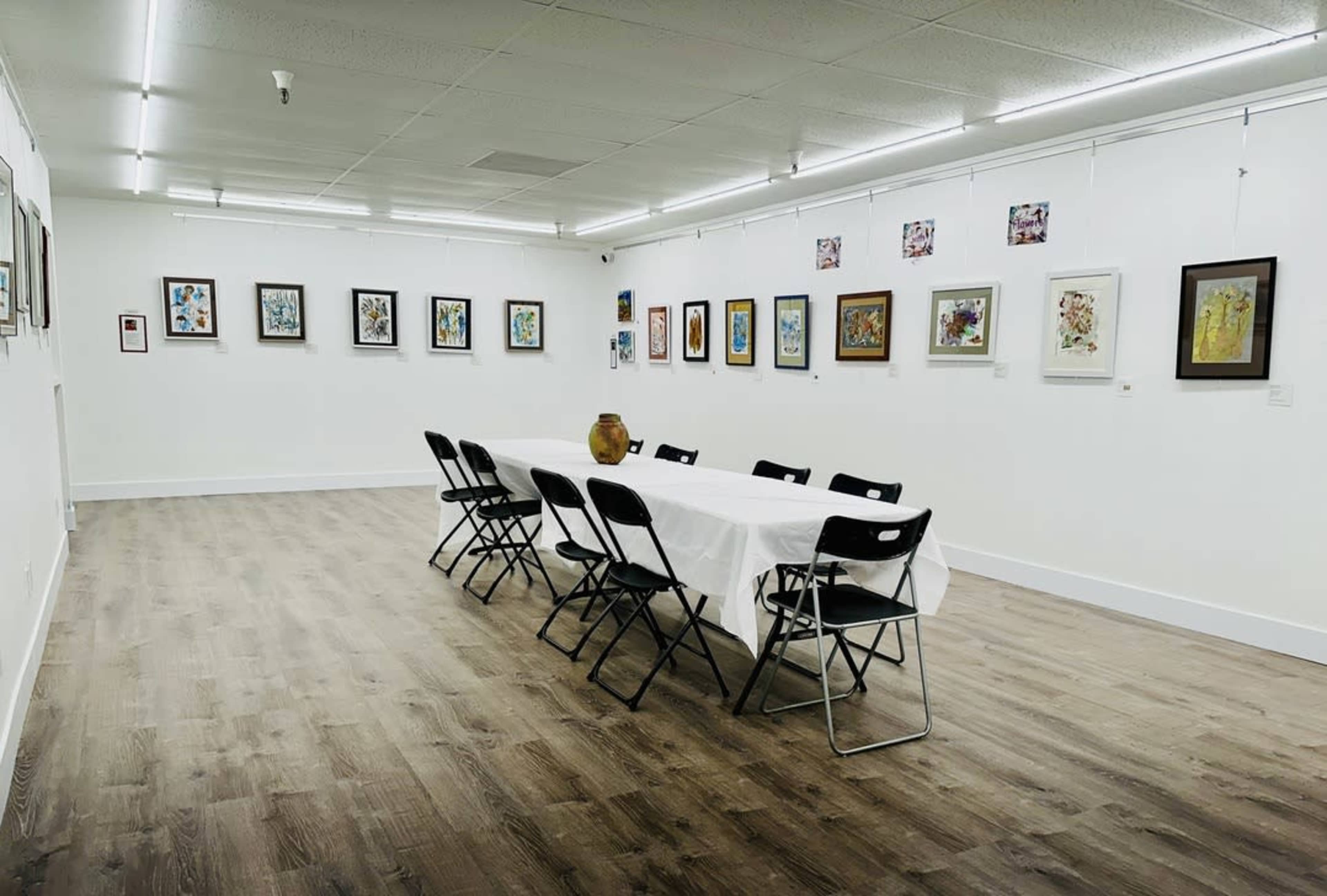 An art gallery with framed paintings on the walls and a long table set with white tablecloth and black chairs arranged around it.