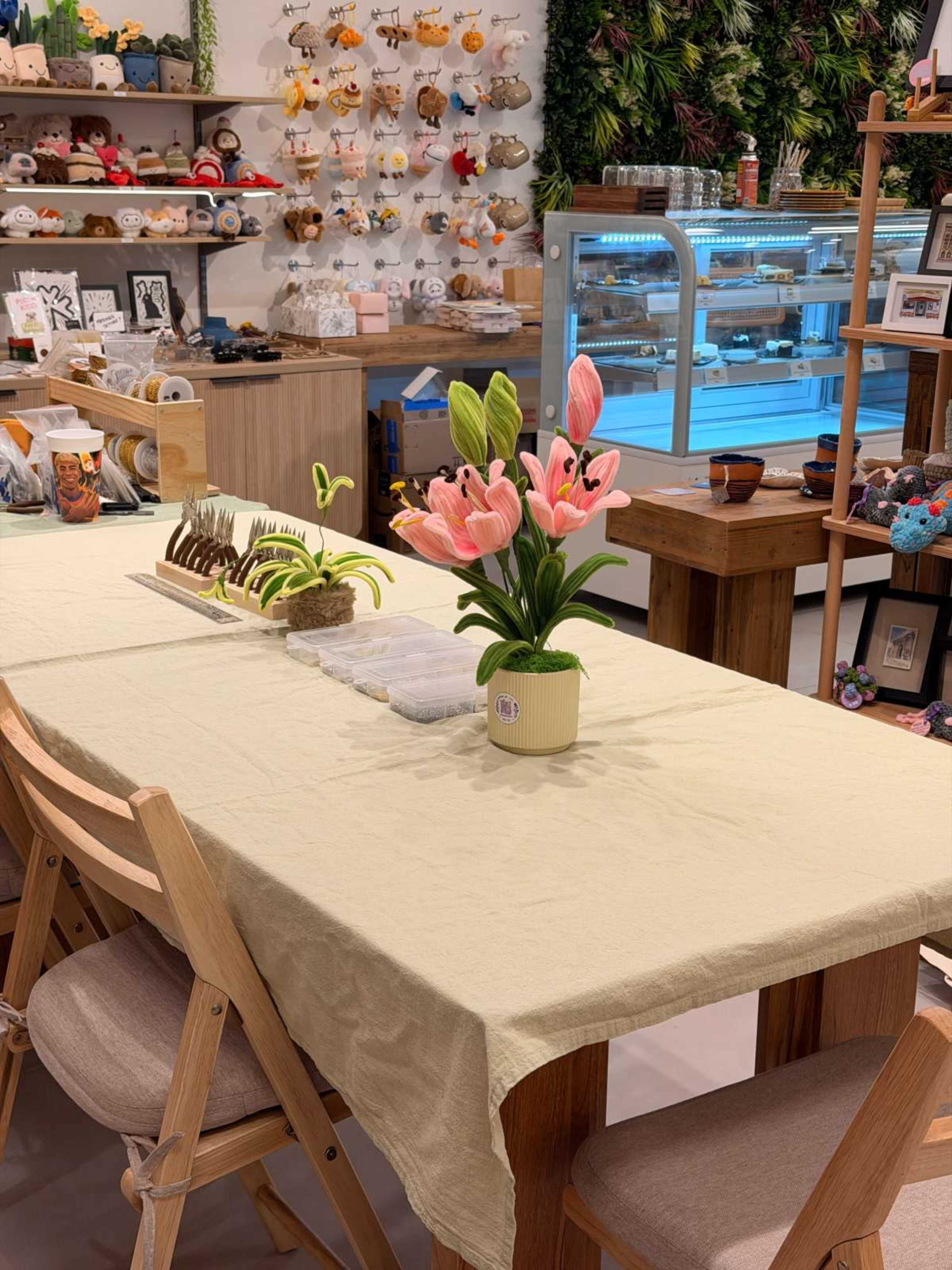 A wooden table is set with a light cloth and features a bouquet of pink flowers at its center, surrounded by a display of various crafts and a glass display case with items in the background.