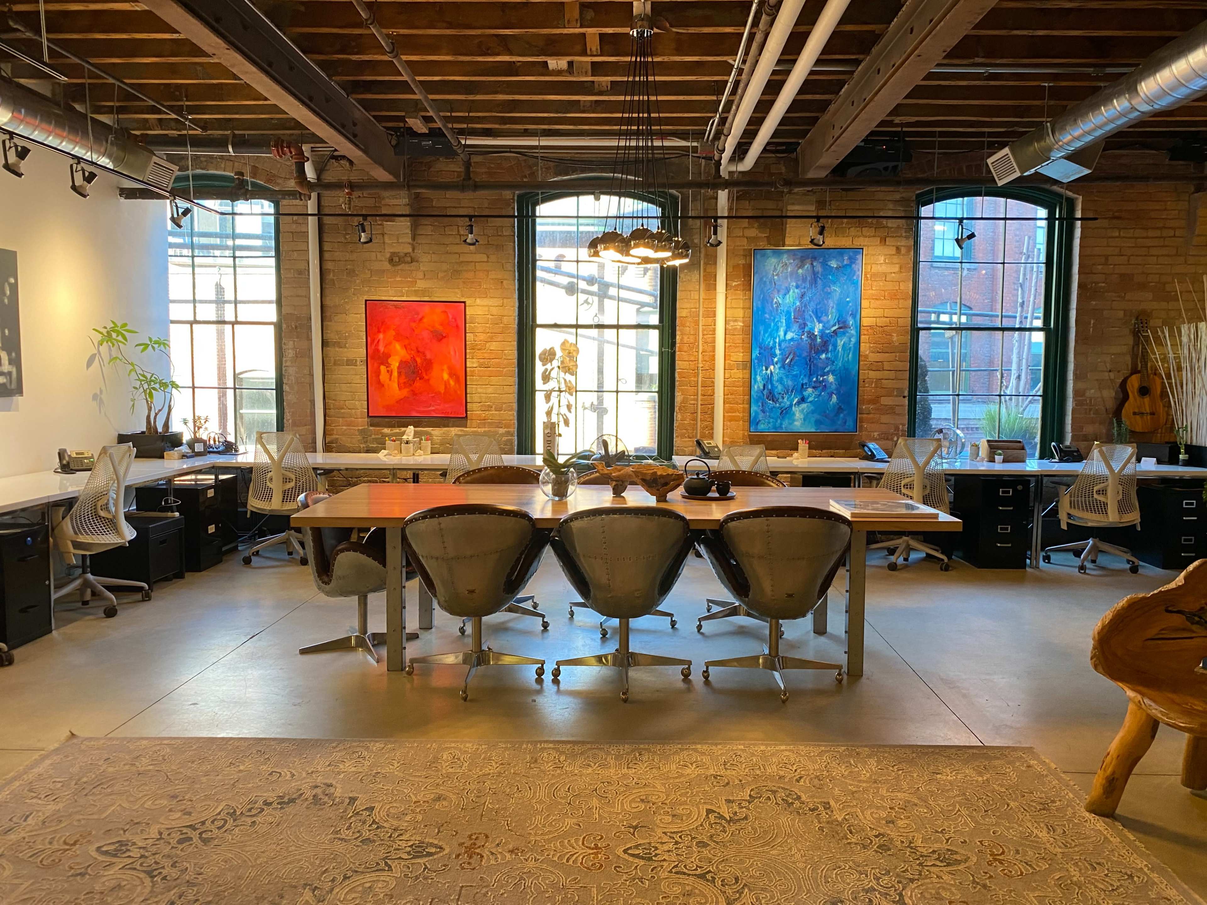 A spacious office with large windows, featuring a long wooden table surrounded by modern chairs and colorful abstract paintings on the walls.