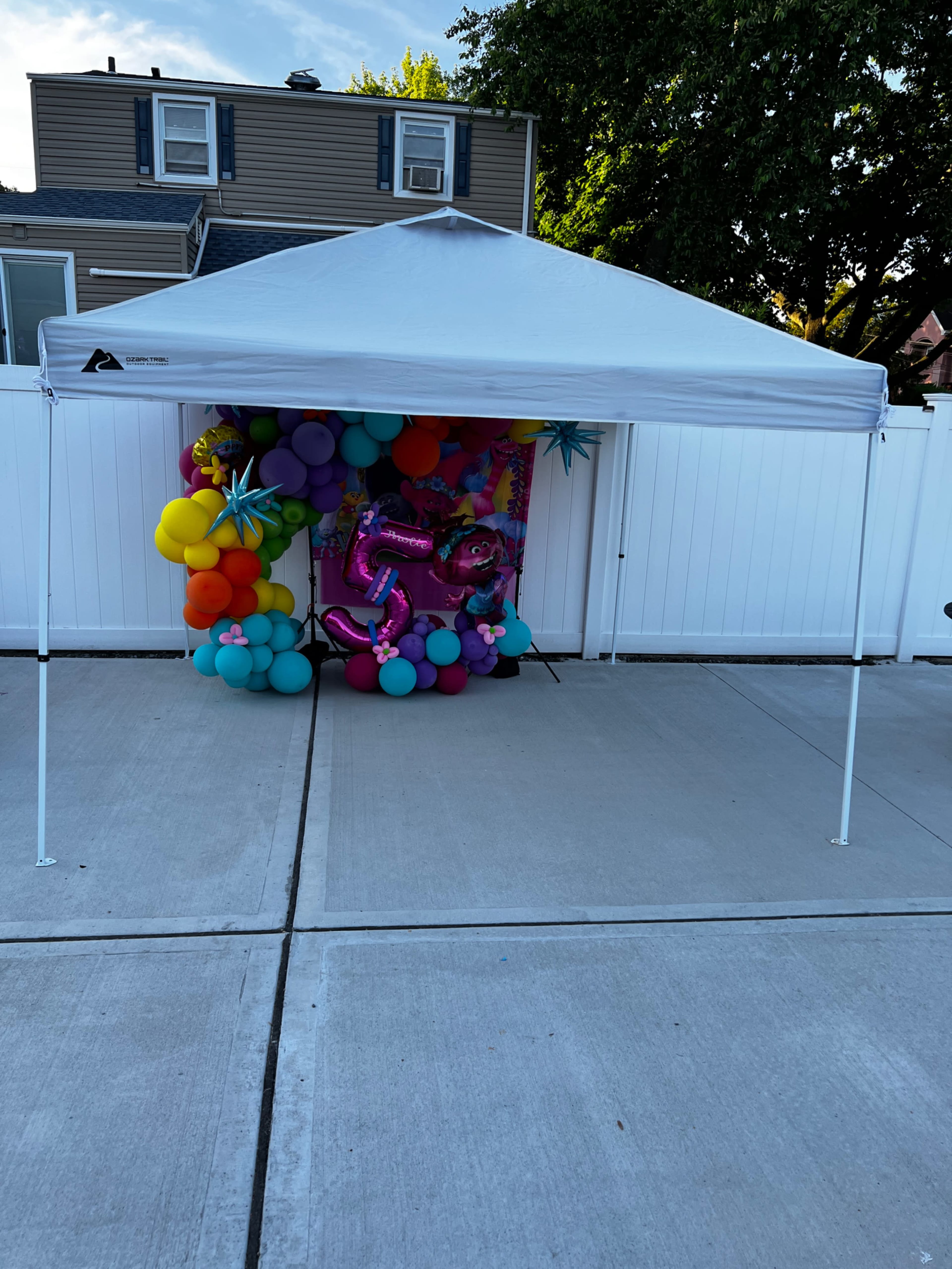 A white canopy tent stands on a concrete surface, with a colorful balloon arrangement and a festive backdrop visible underneath.