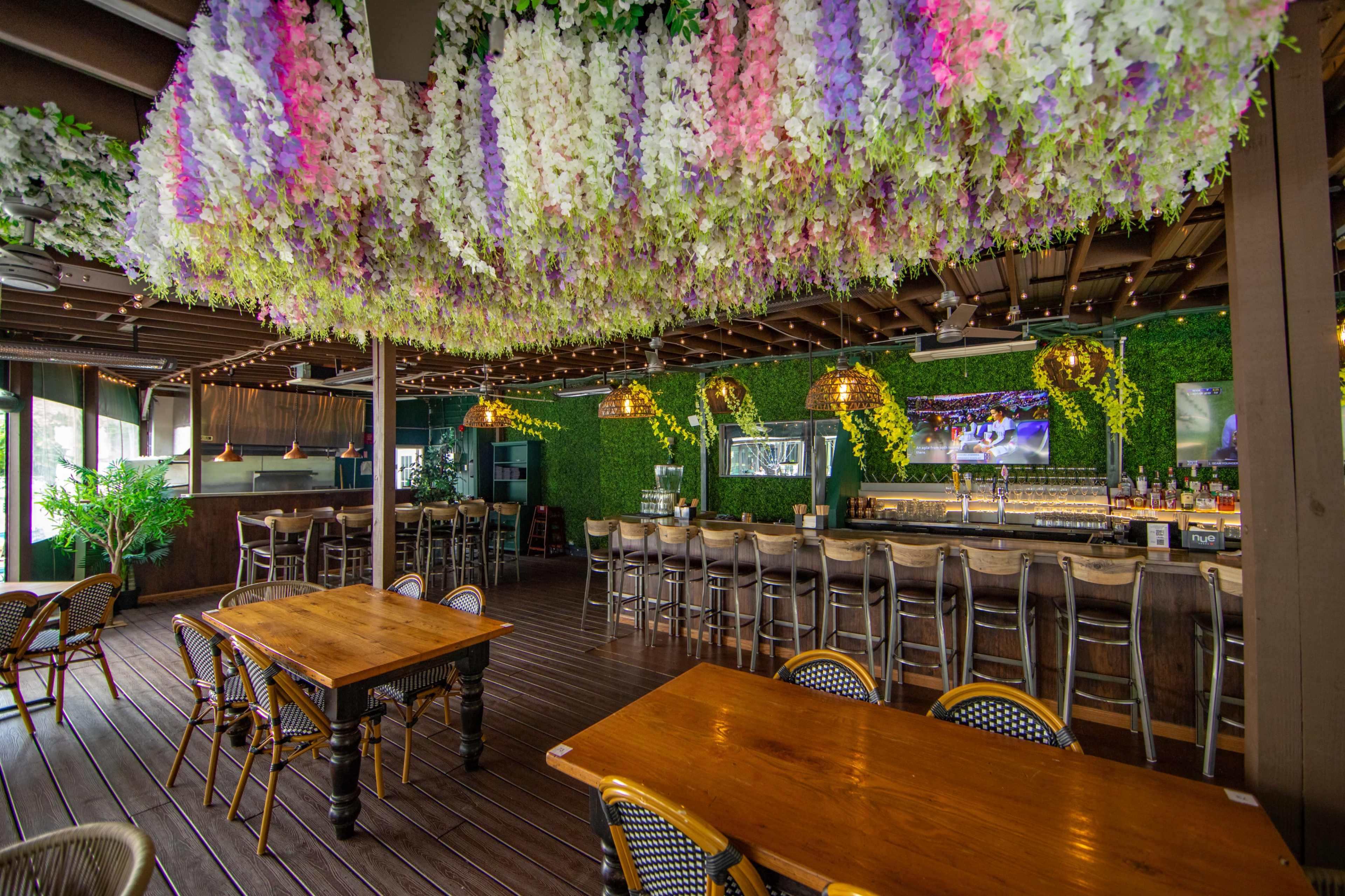 The image shows a bar interior featuring wooden tables, a long counter with stools, and a ceiling adorned with decorative floral arrangements.