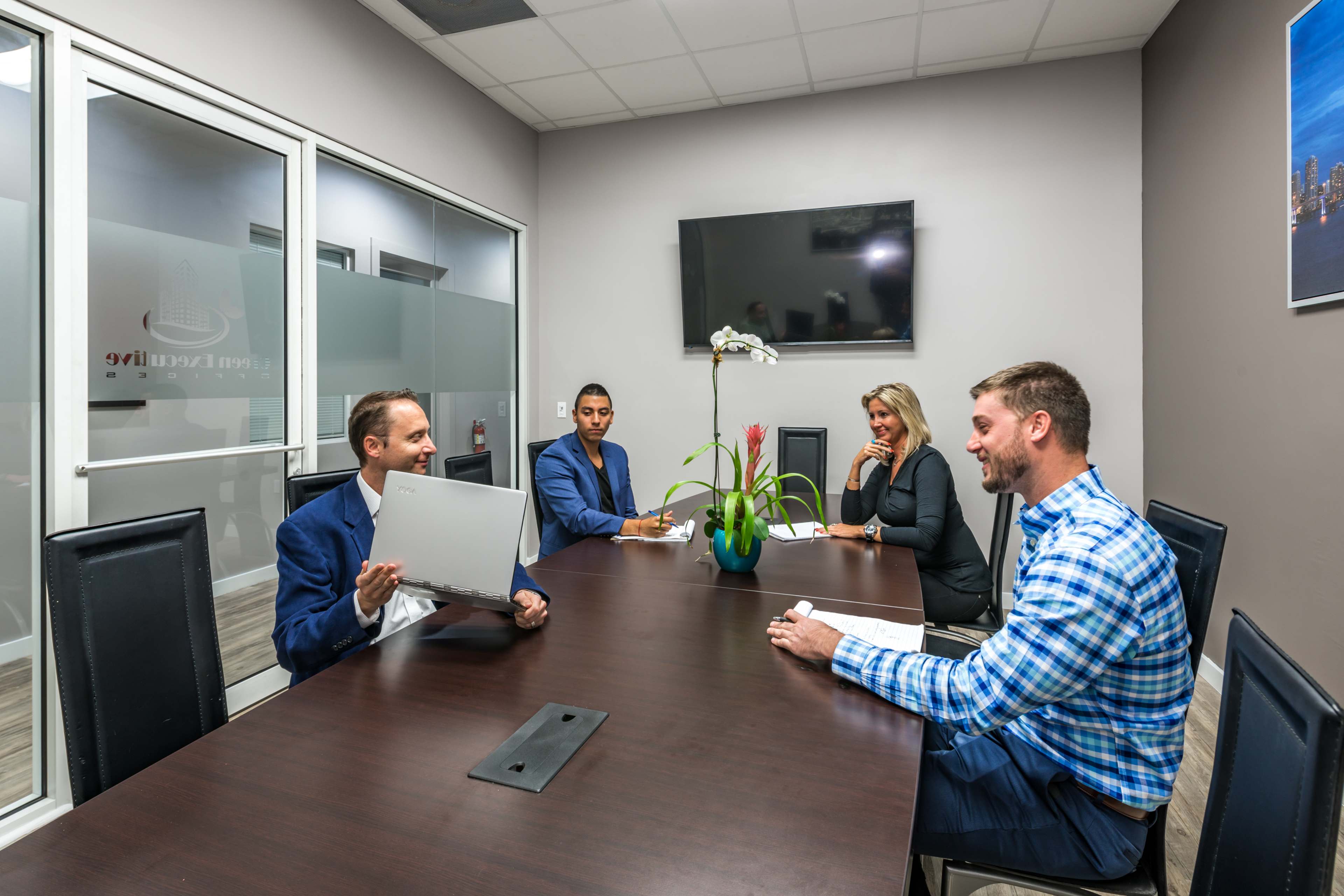 A group of four professionals is seated around a conference table in a modern meeting room, with one person holding a laptop and discussing.