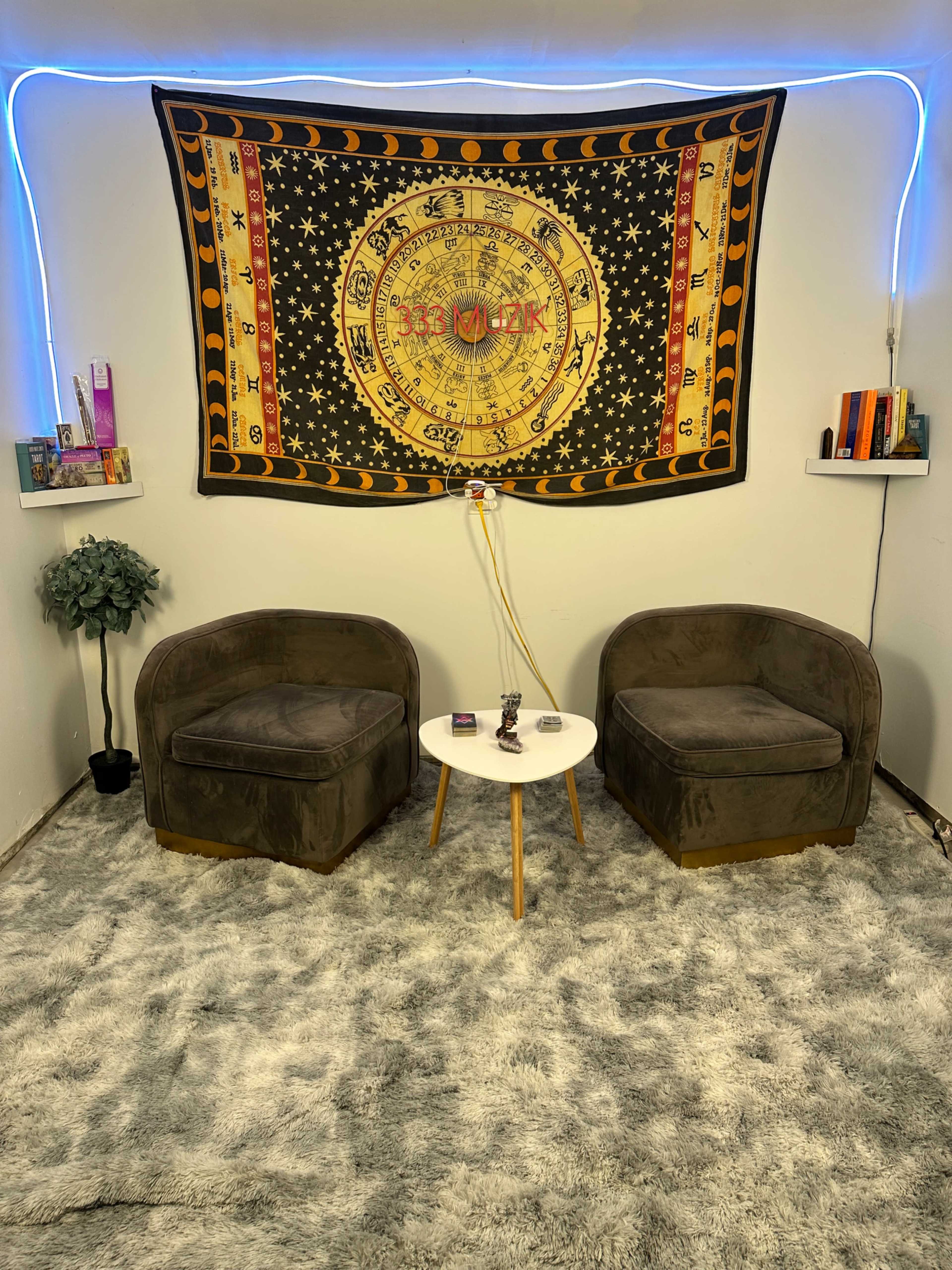 The image shows a cozy room setup with two brown chairs facing a small white table, decorated with a tapestry featuring astrological symbols hanging on the wall, and shelves with books and a plant.