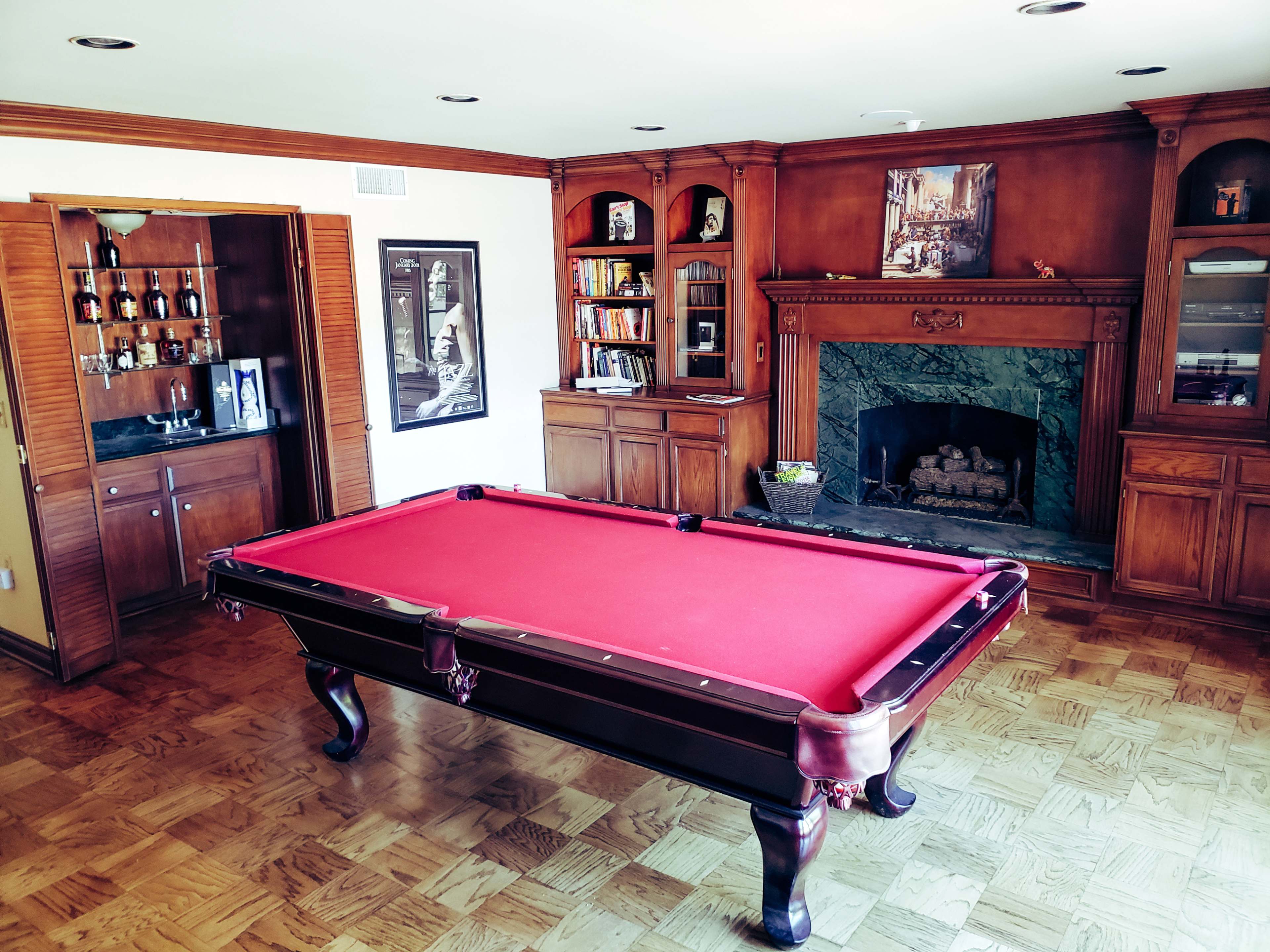 A billiard table with a red cloth is centered in a room featuring wooden cabinetry, a fireplace, and shelves filled with books and decorative items.