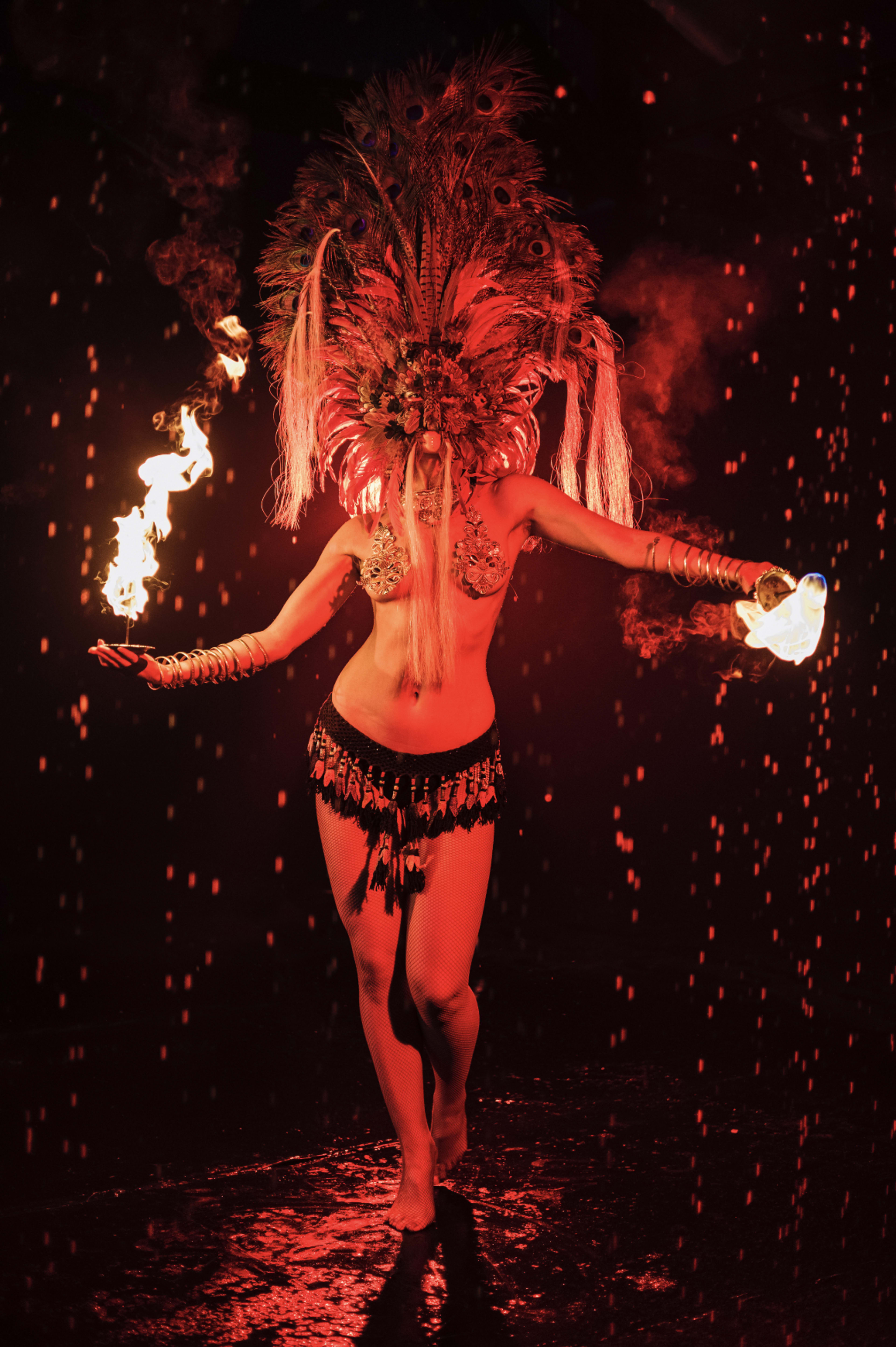 A performer with a decorative feather headdress and a tribal outfit executes a fire dance in a dark environment with red lighting.
