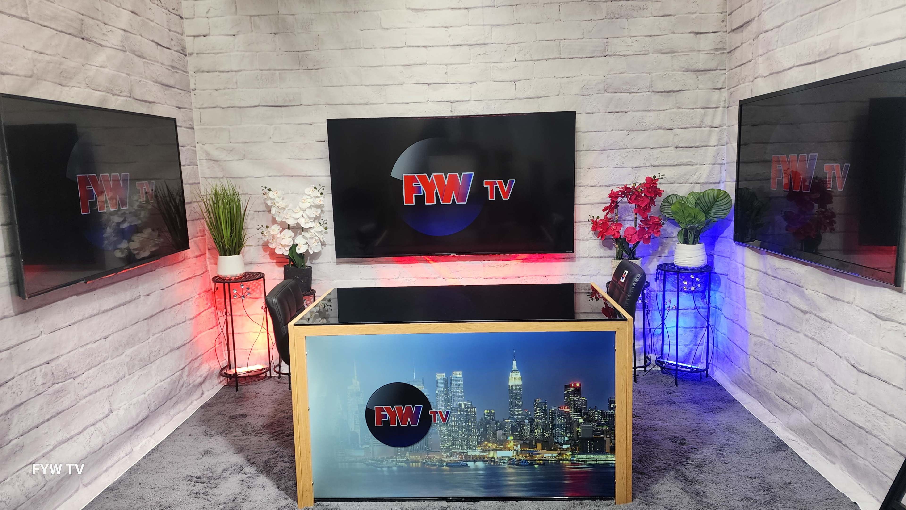 The image shows a television studio setup with a central glass table and multiple monitors displaying the logo "FWY TV" against a backdrop of a city skyline.