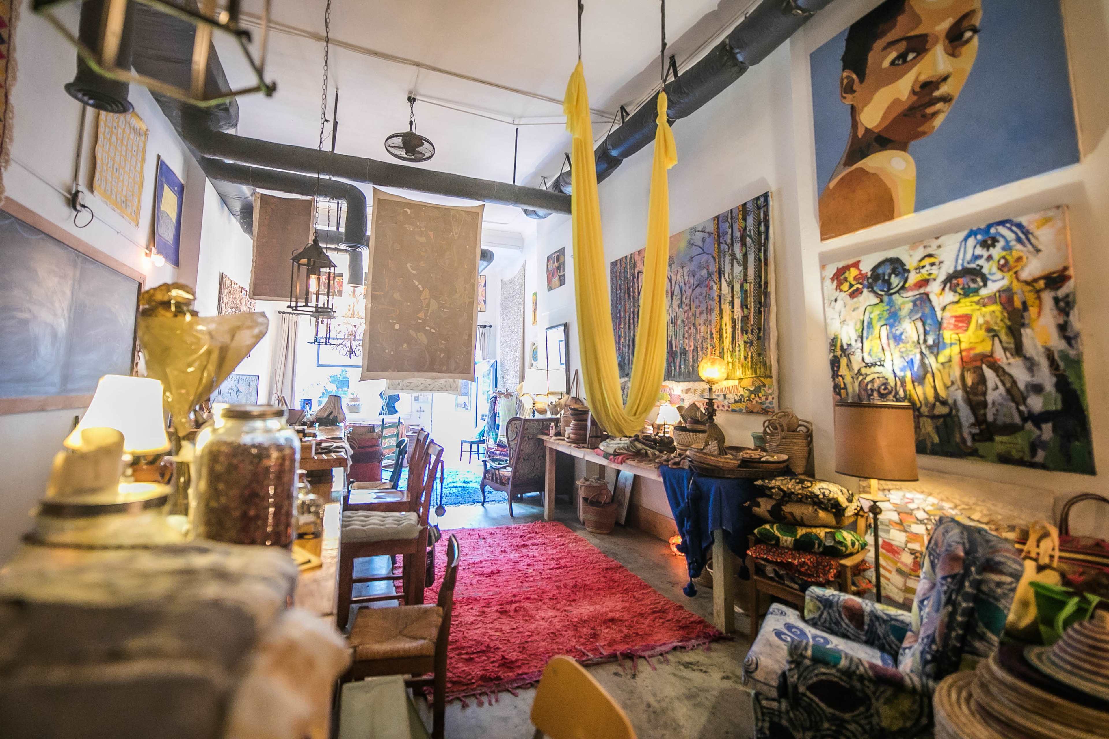 An eclectic art-filled interior of a cozy cafe or shop, featuring colorful paintings, textiles, and various decorative items arranged throughout the space.