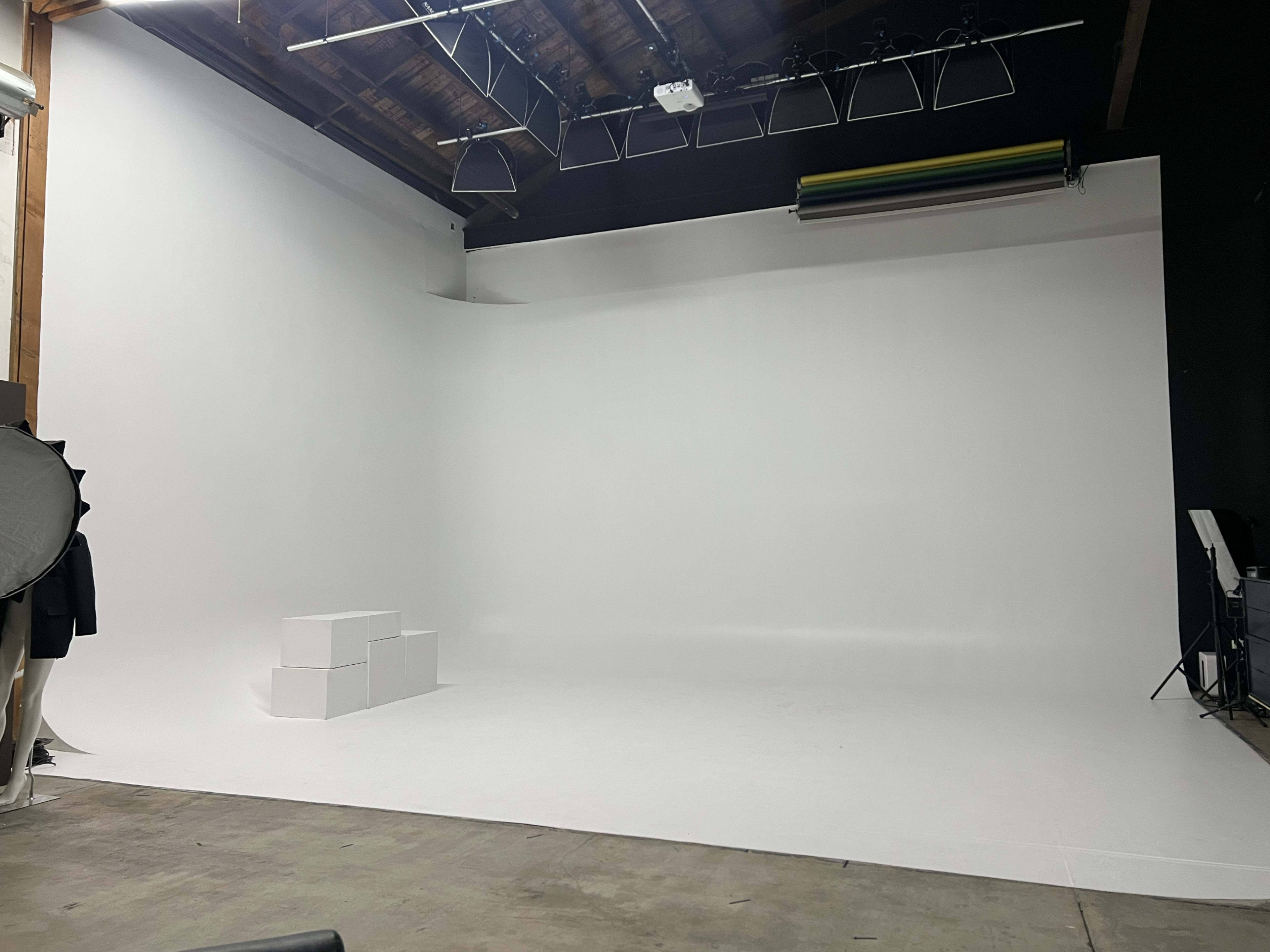 The image shows a spacious photo studio with a white backdrop, minimal props, and soft lighting equipment.