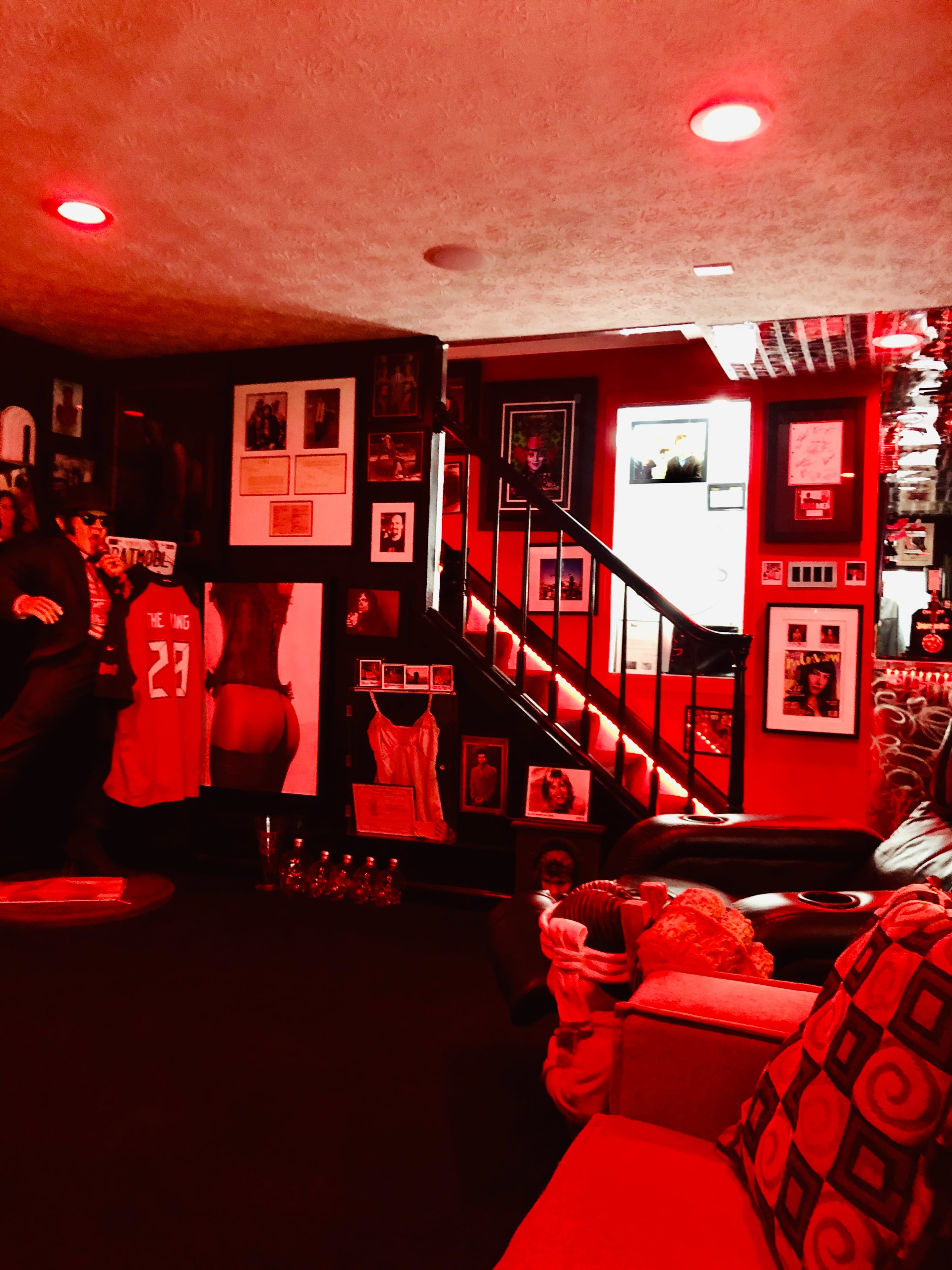 The image shows a dimly lit room adorned with framed photographs and artwork, featuring a staircase at the back and red lighting throughout the space.