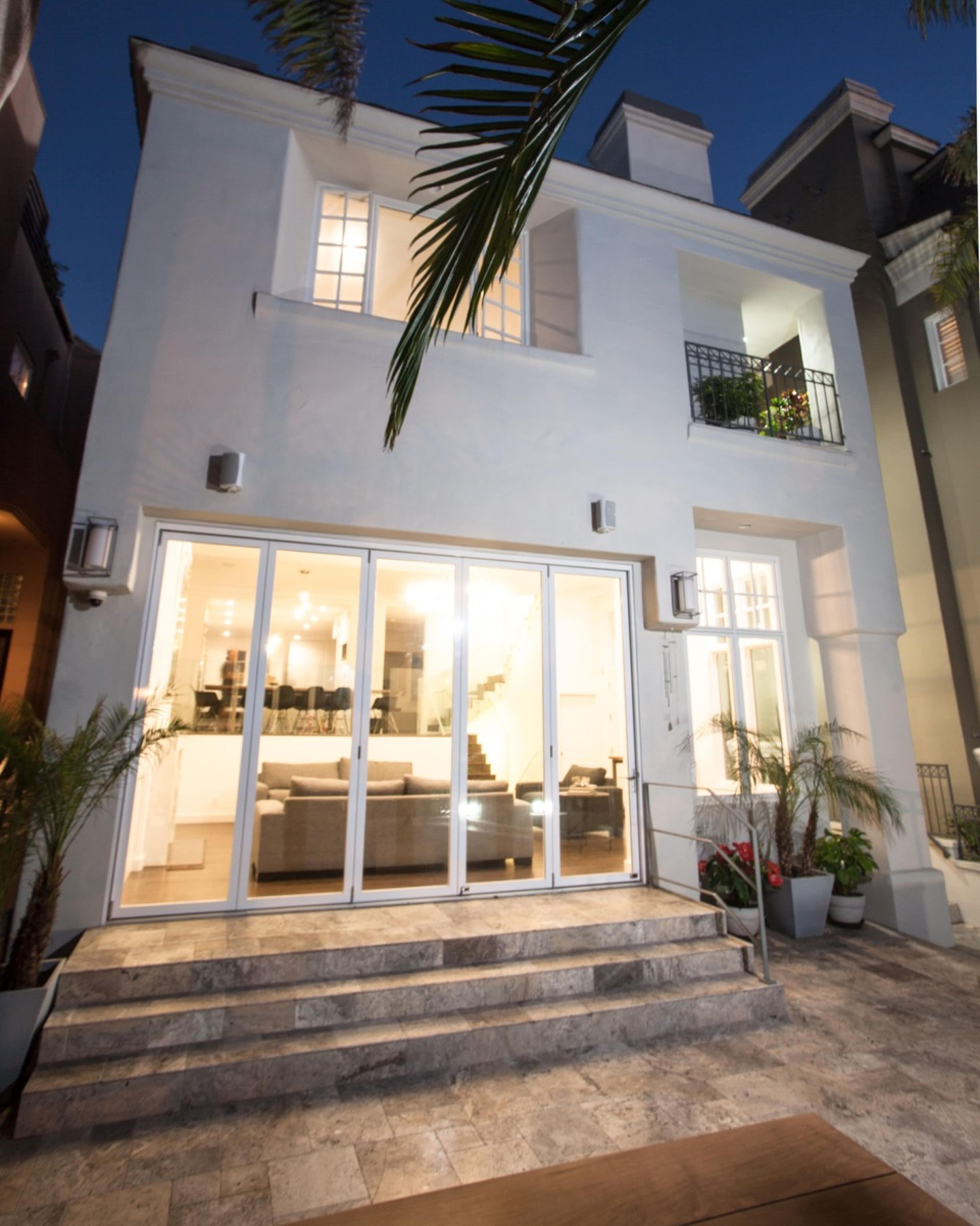 A modern two-story house features large glass doors leading to a well-lit interior, surrounded by potted plants and palm trees.