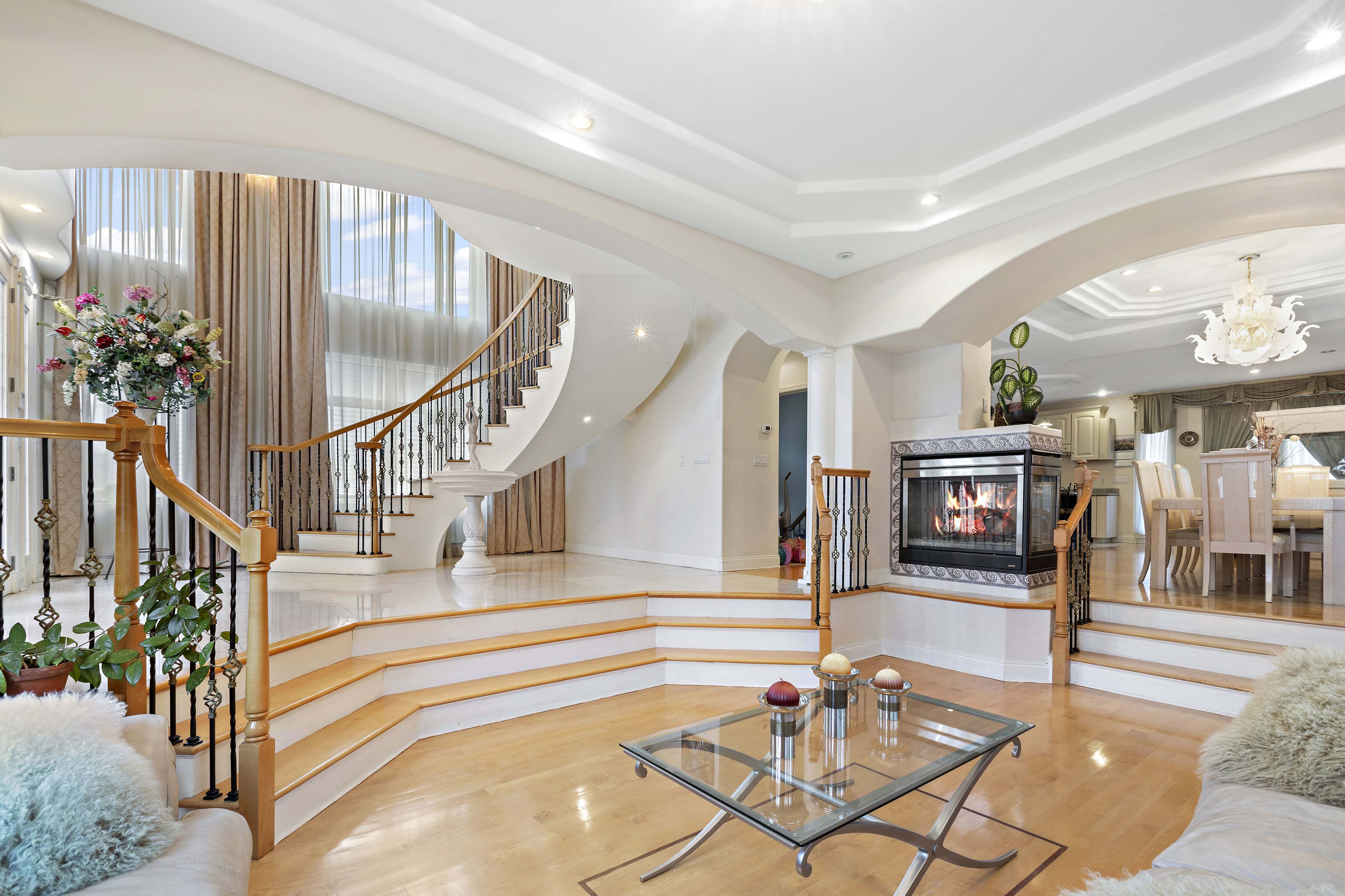 A spacious interior with a curved staircase, a modern fireplace, and large windows allowing light to enter.