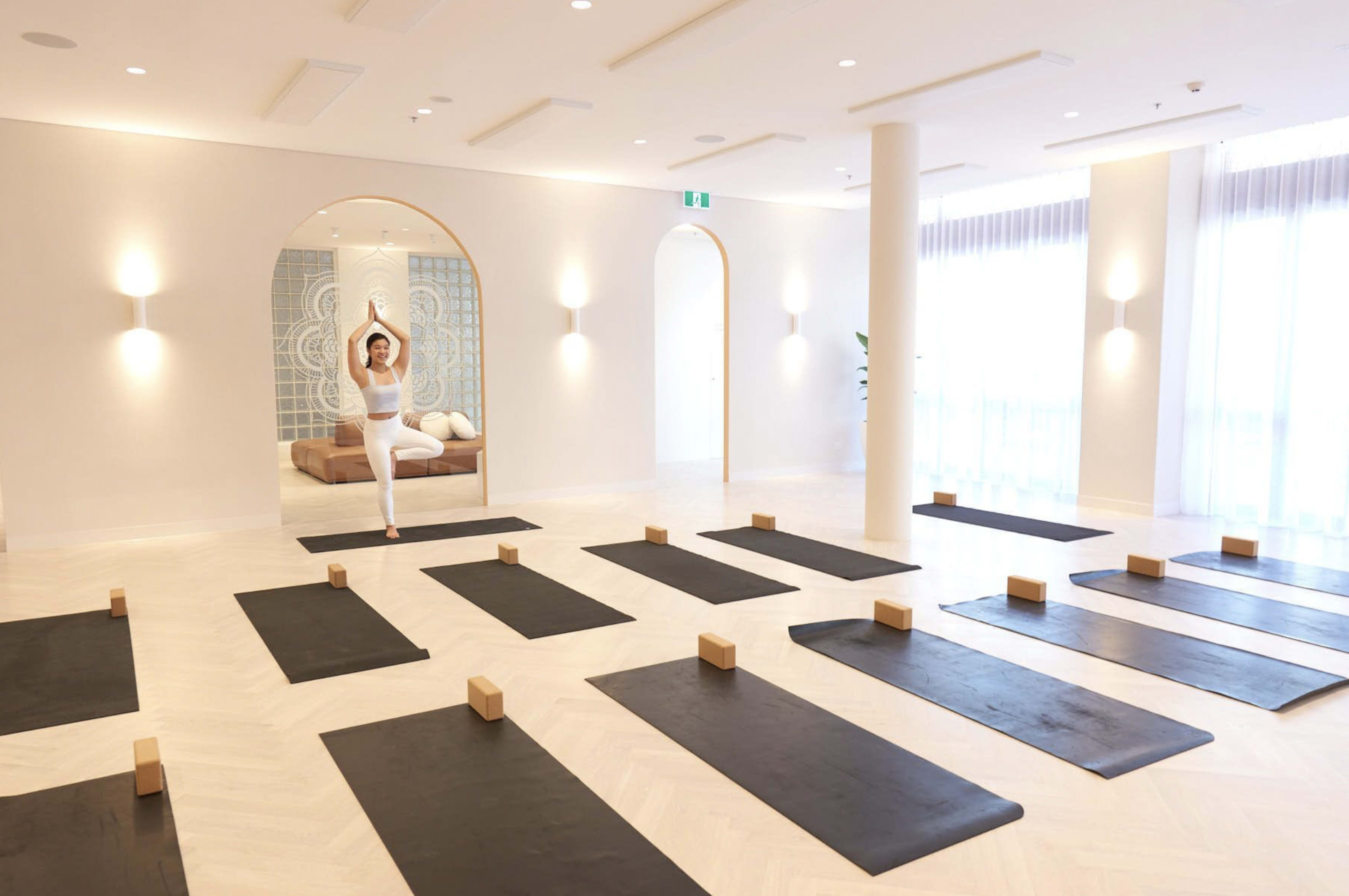 A yoga studio features neatly arranged mats with wooden blocks, and a person practicing a pose in the background.