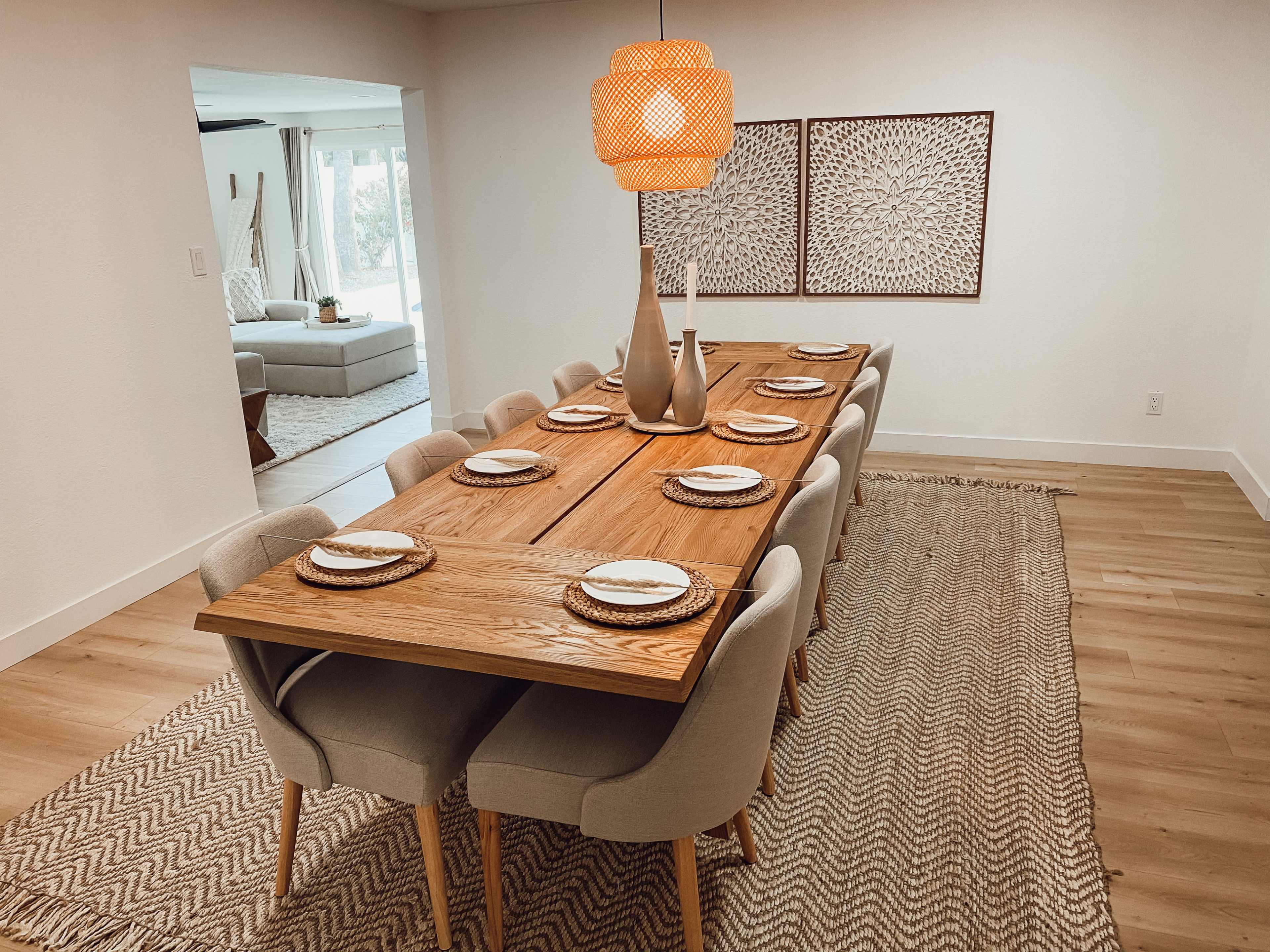 A long wooden dining table is set with plates and utensils, surrounded by upholstered chairs on a woven rug, with a decorative lamp and art pieces in the background.