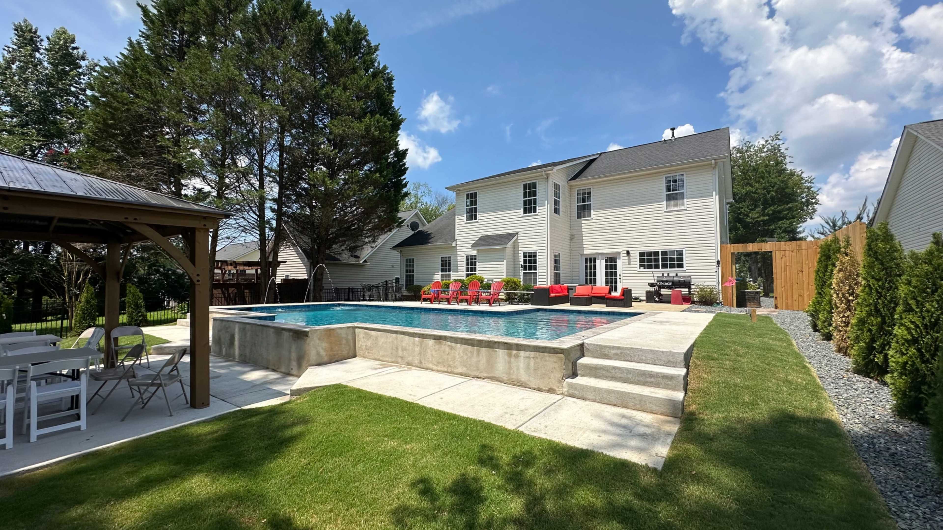 The image shows a rectangular in-ground swimming pool with lounge chairs around it, adjacent to a modern two-story house and surrounded by green grass and trees.