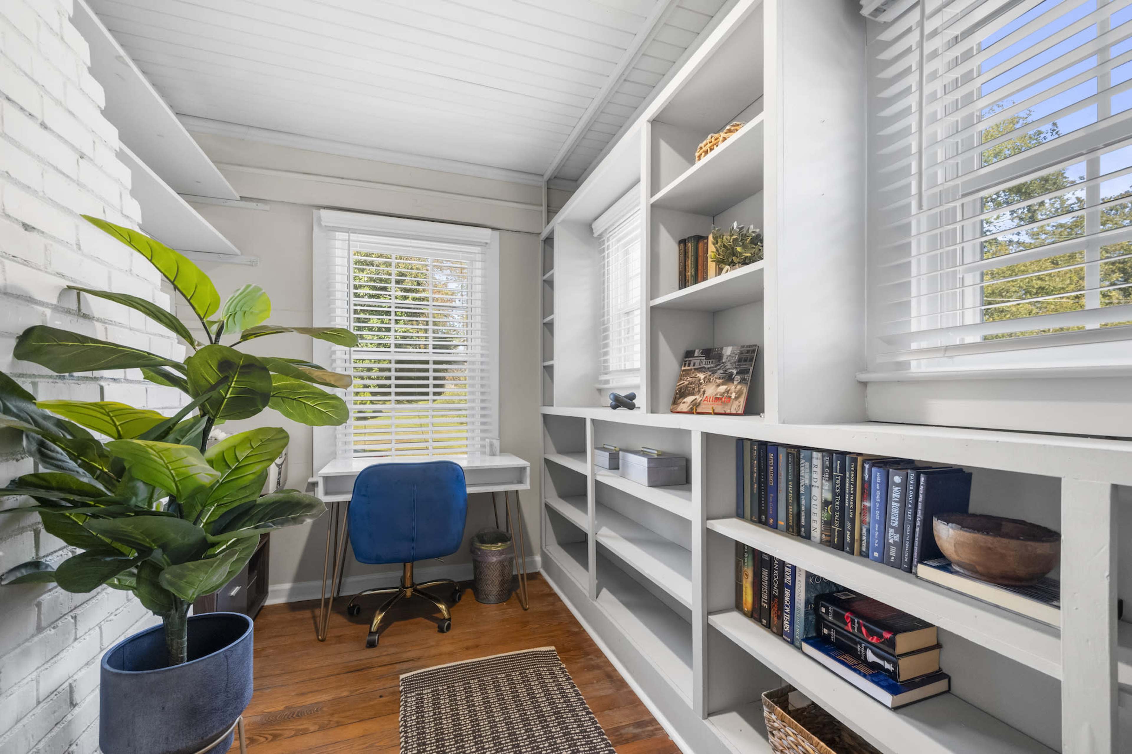 A small home office features built-in shelves filled with books, a blue chair, and a desk by a window with white blinds, alongside a potted plant.