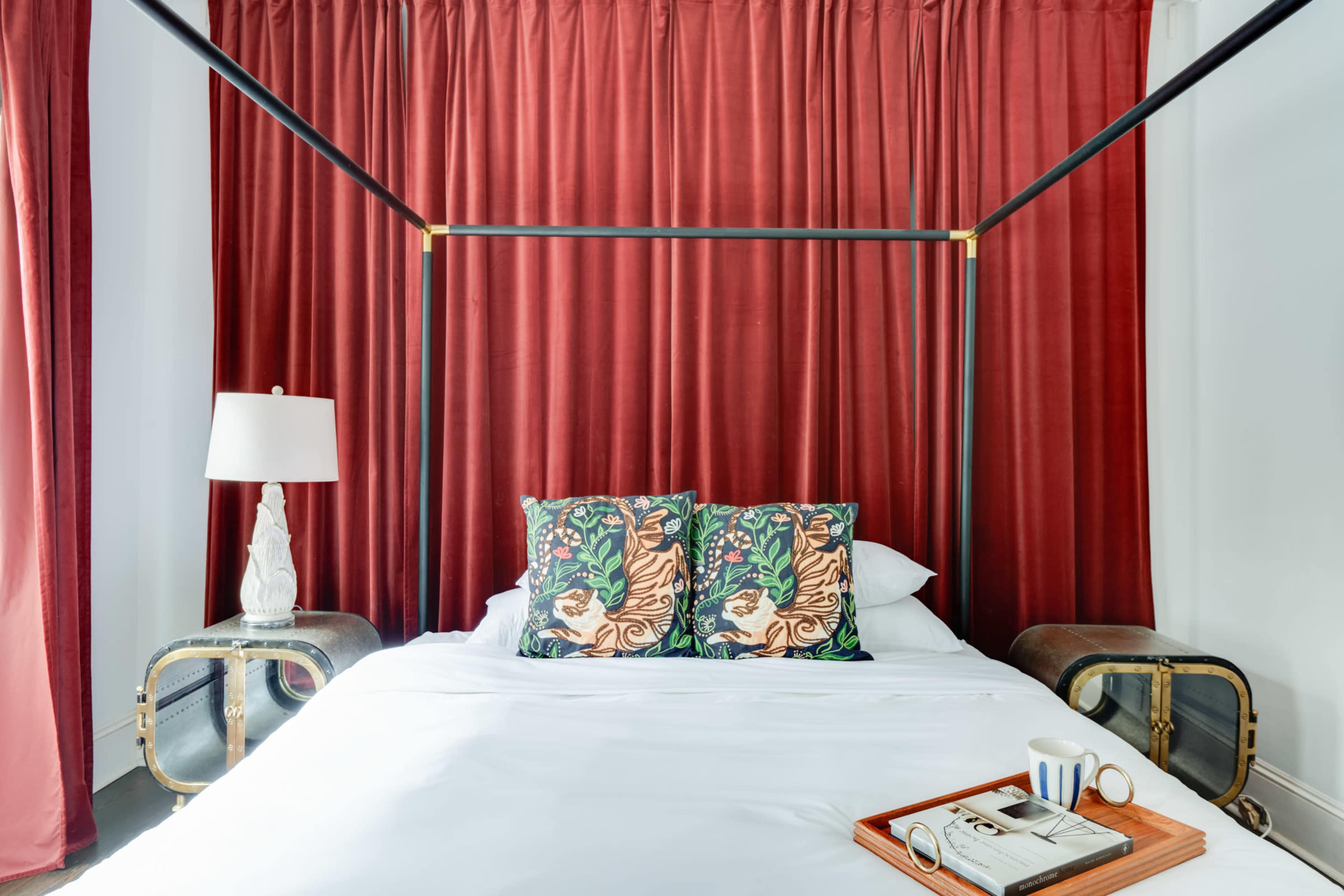 The image shows a four-poster bed with decorative pillows, set against a backdrop of red curtains.
