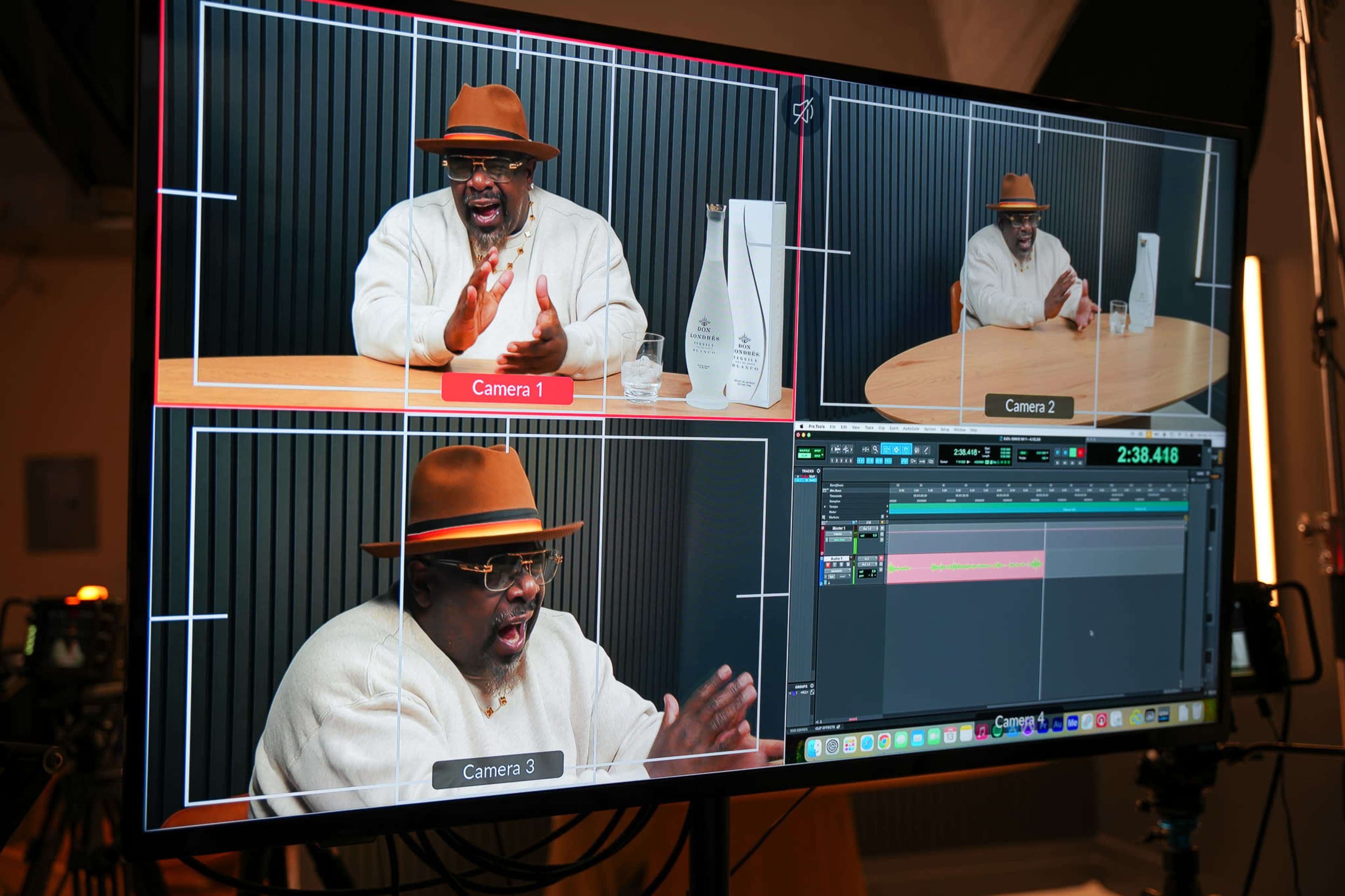 The image shows a monitor displaying a multi-camera setup, featuring a man in a hat speaking animatedly in front of a microphone and a wooden table.