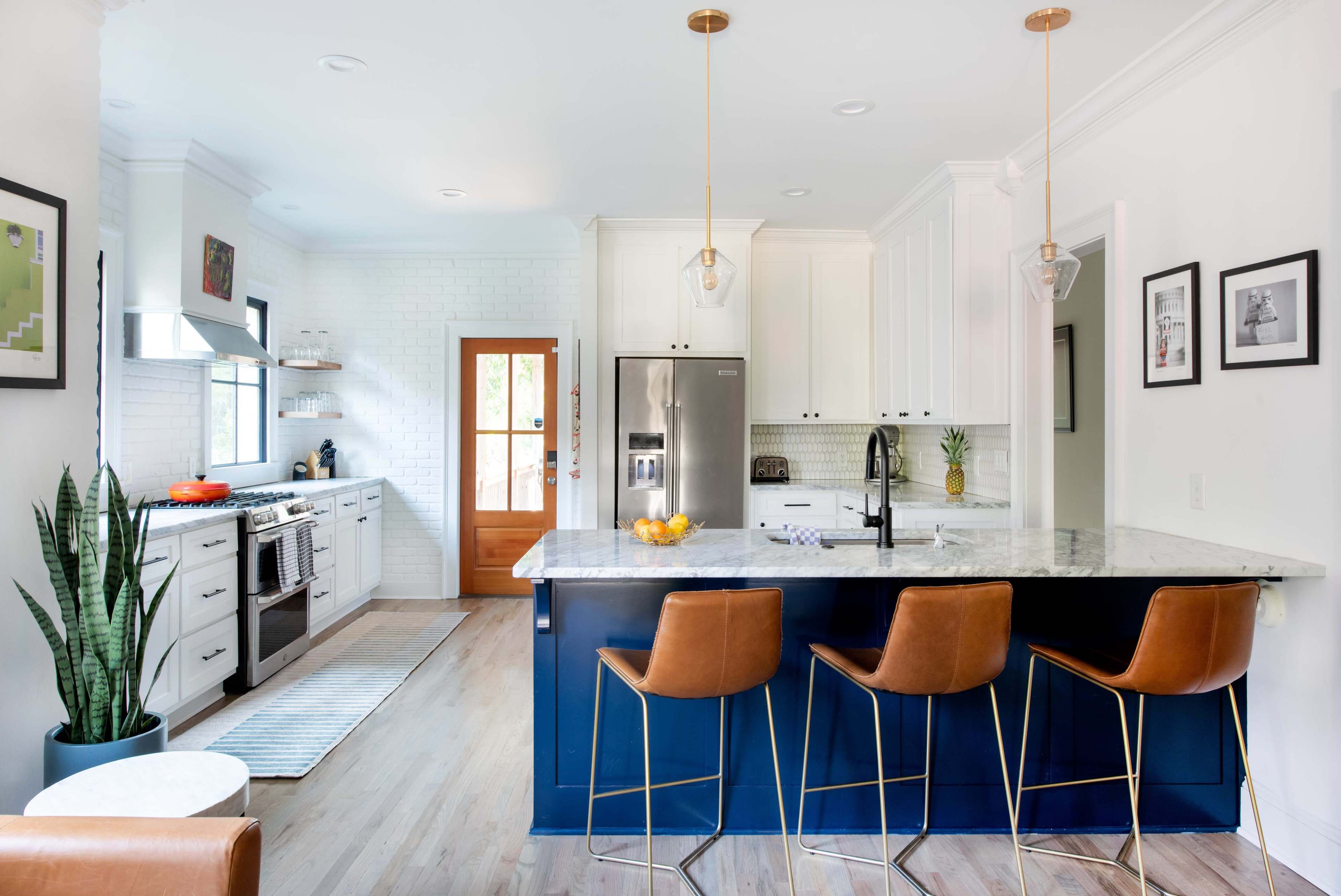The image shows a modern kitchen with white cabinetry, stainless steel appliances, a marble countertop, and a large island with three brown bar stools.