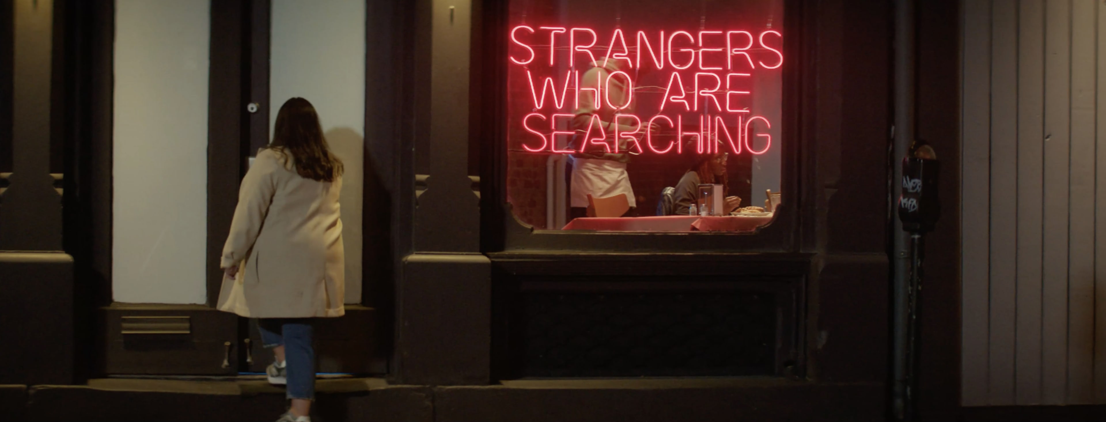 A woman approaches a storefront with a neon sign that reads "STRANGERS WHO ARE SEARCHING."