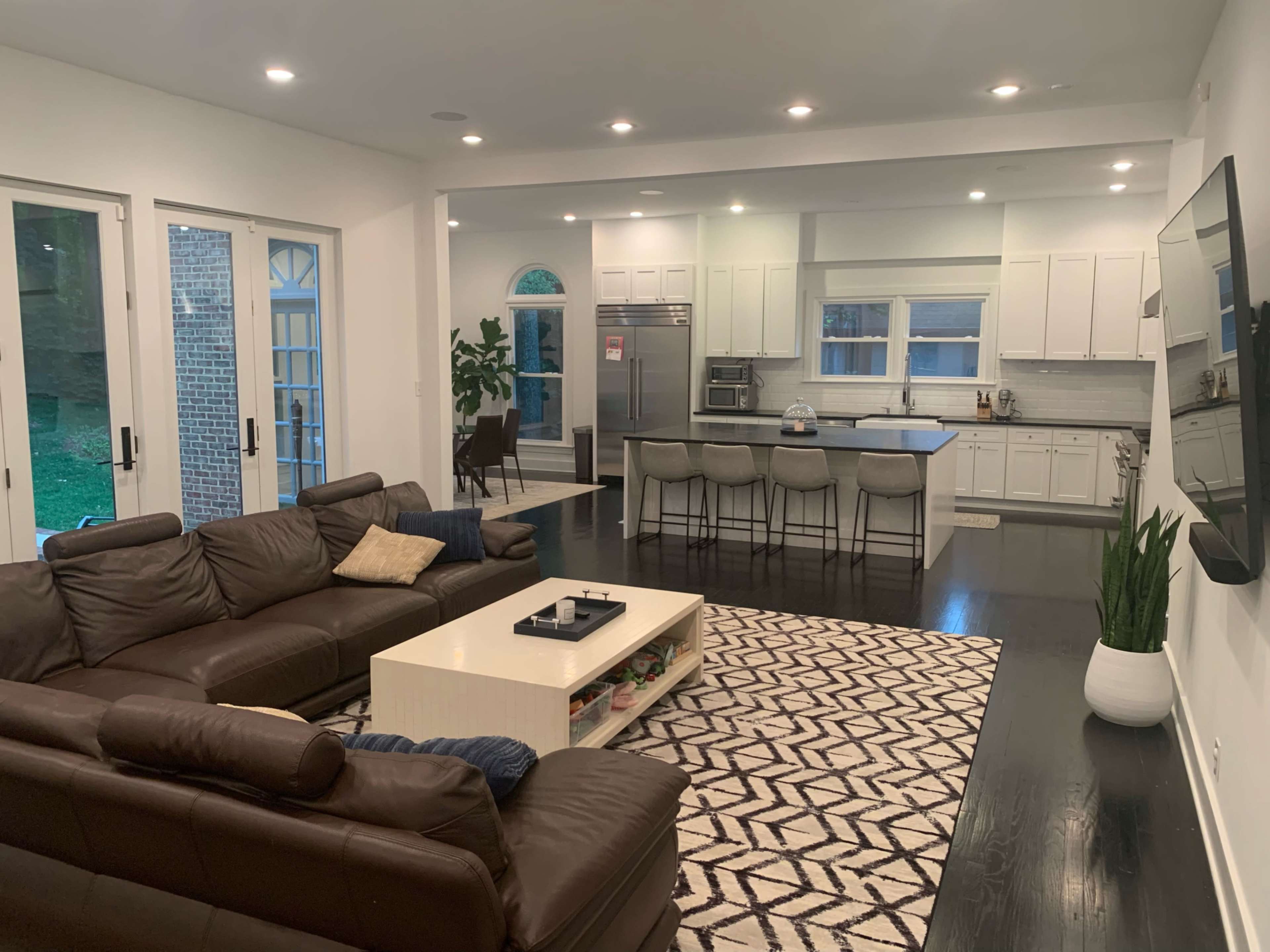 A modern living room with a brown leather sofa, a white coffee table, and a kitchen area featuring white cabinets and a large island with bar stools.