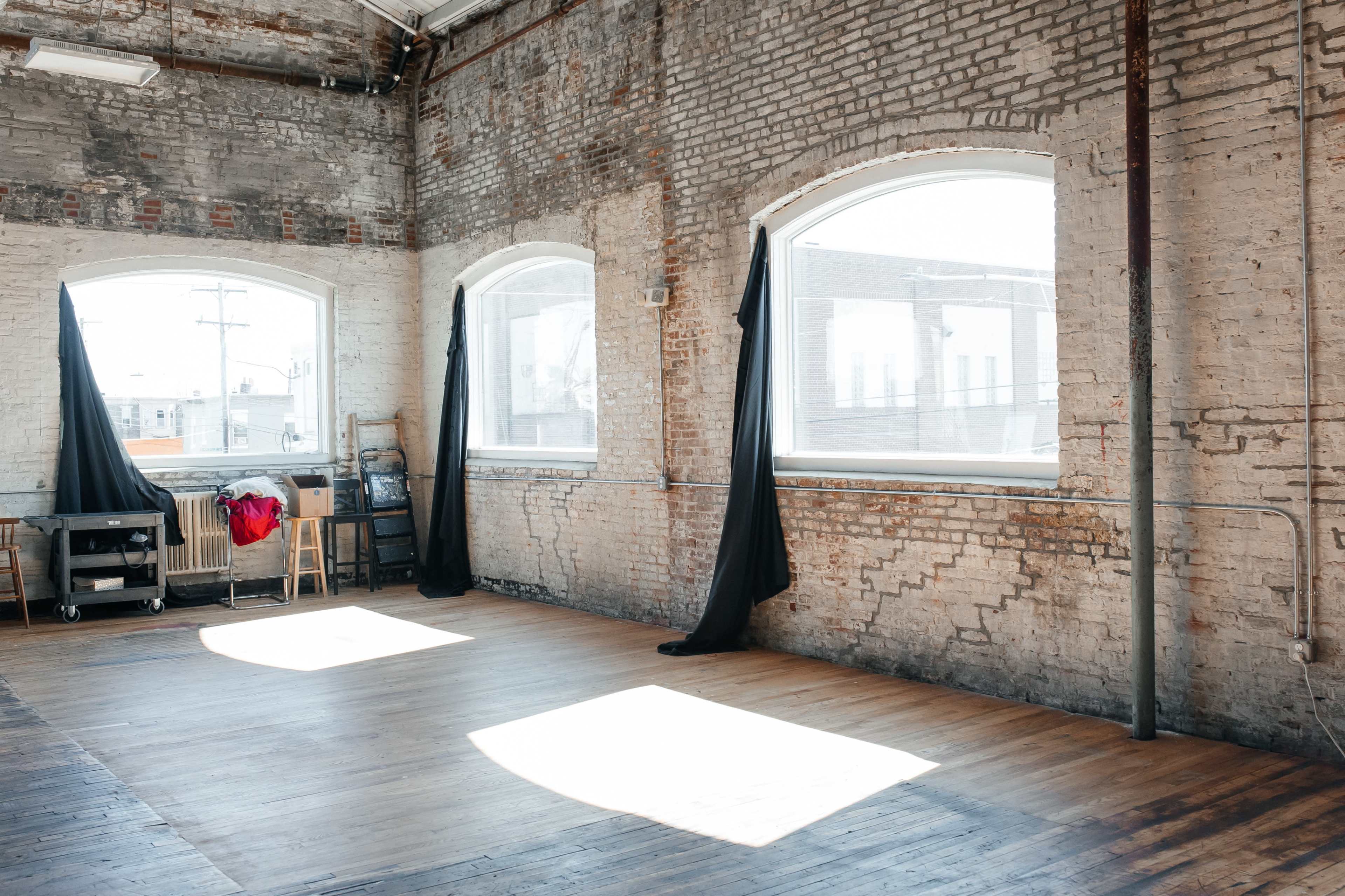 An empty room with exposed brick walls and large windows, featuring wooden flooring and two black curtains drawn back.