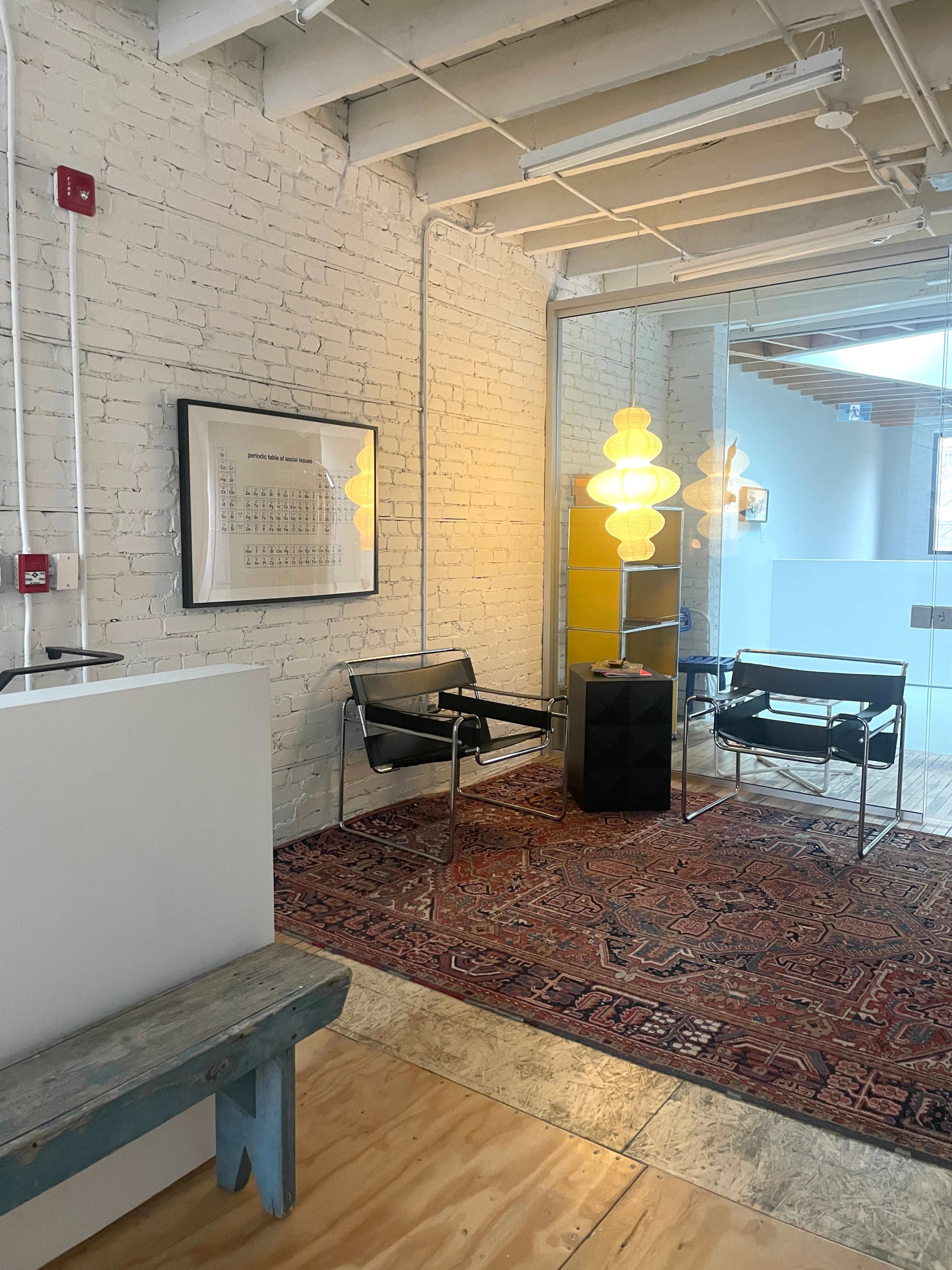 The image shows a modern interior space with a wooden floor, featuring two black chairs, a large area rug, and a brightly illuminated light fixture near a white brick wall.