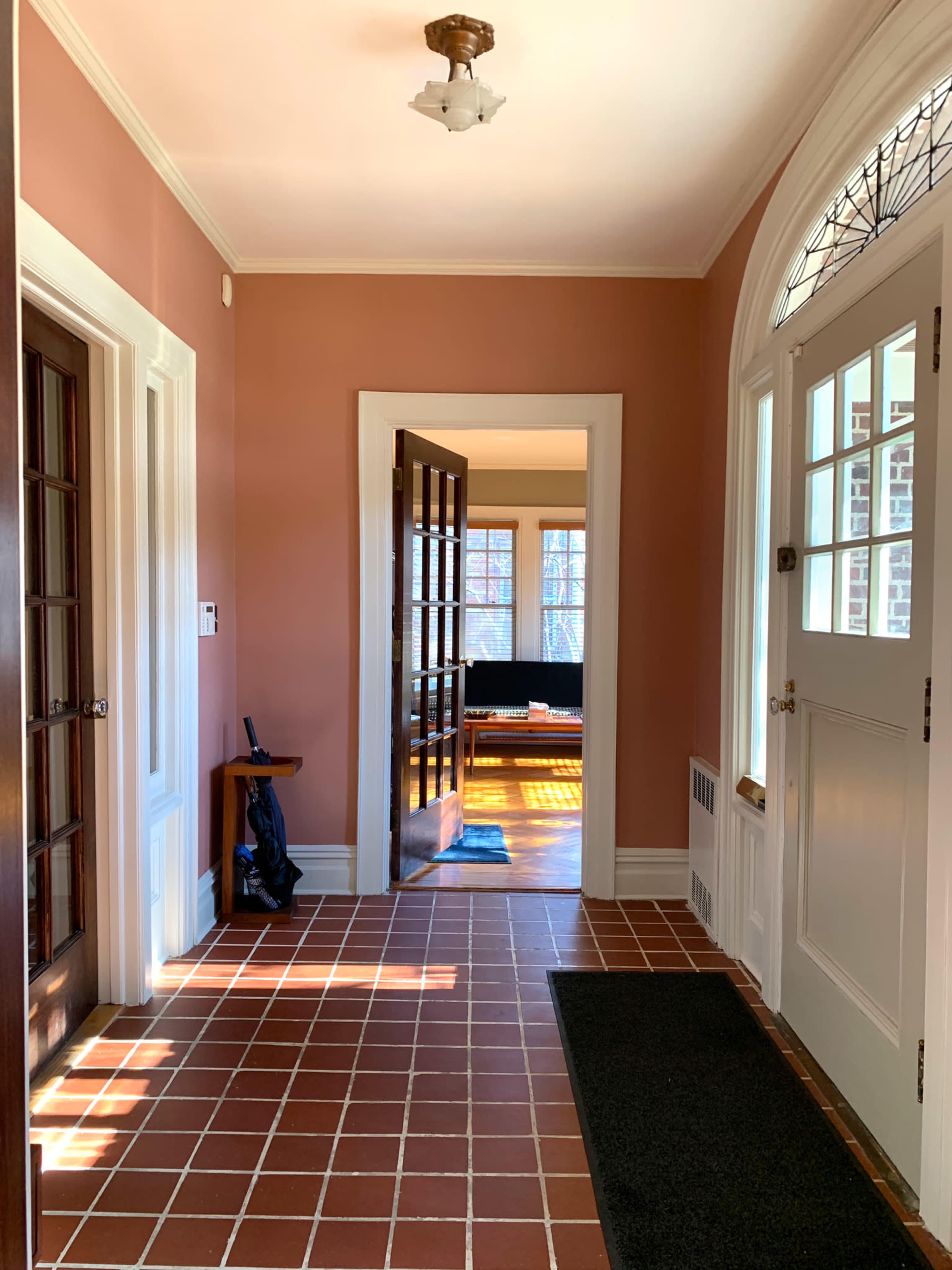 A hallway with tiled flooring, two wooden doors on the left, and an open door at the end leading to a sunlit room.