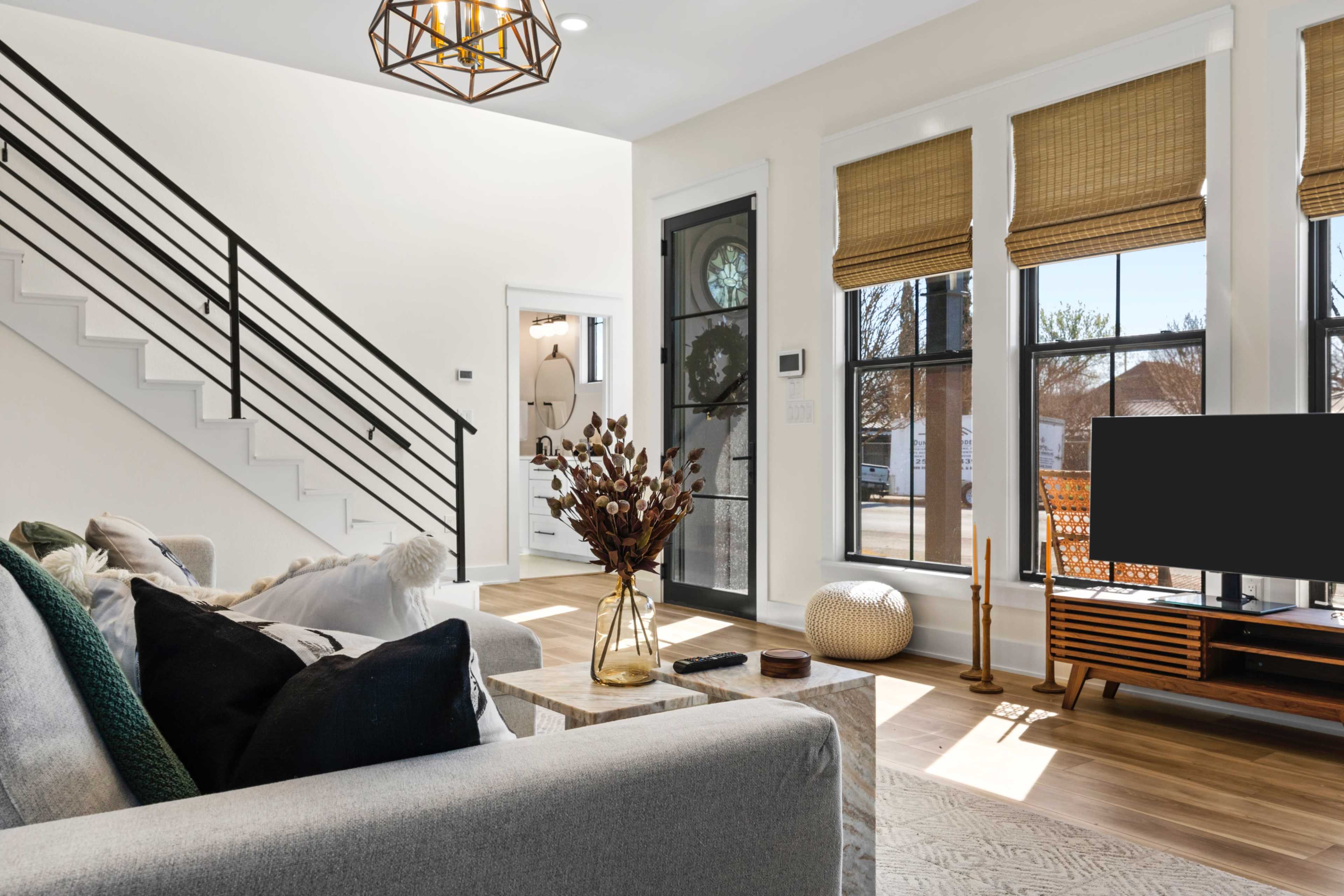 A modern living room with a gray sofa, a coffee table, and large windows featuring woven shades, along with a staircase in the background.