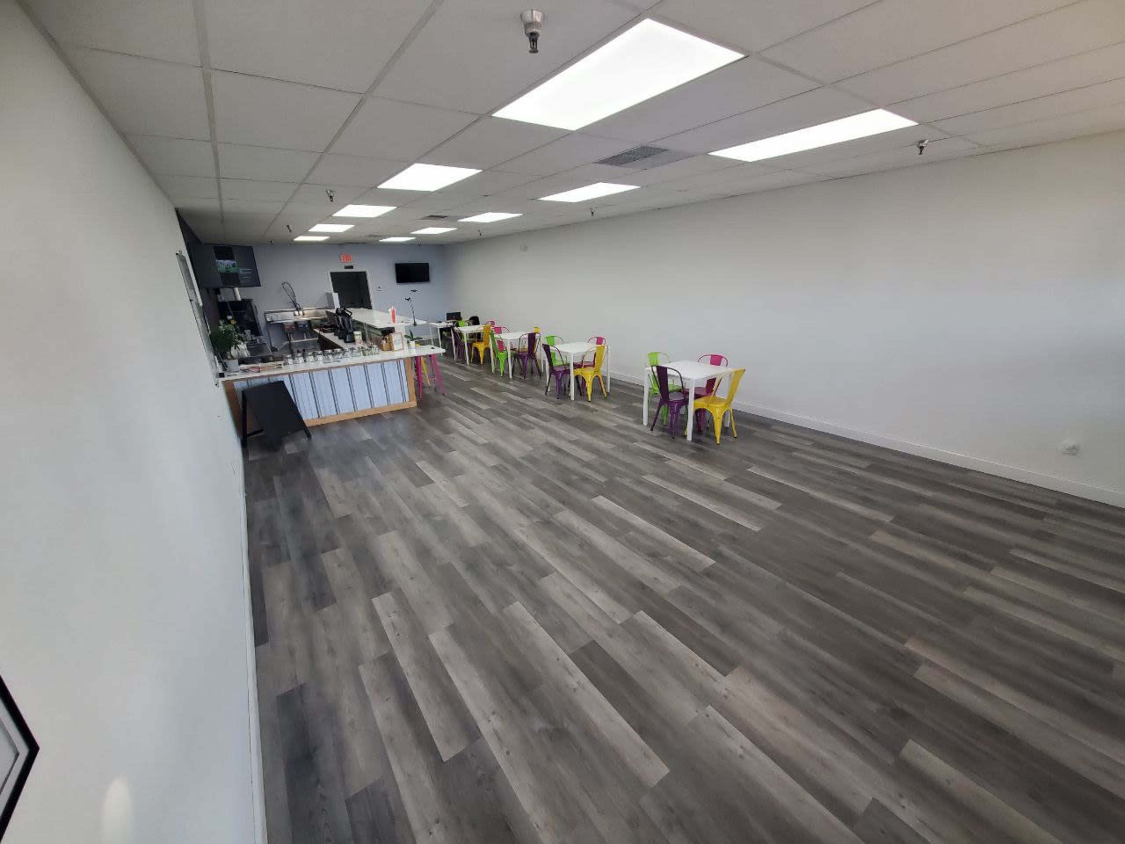 A spacious, empty room with gray wood-like flooring, white walls, colorful chairs arranged at tables along the sides, and a counter at one end.