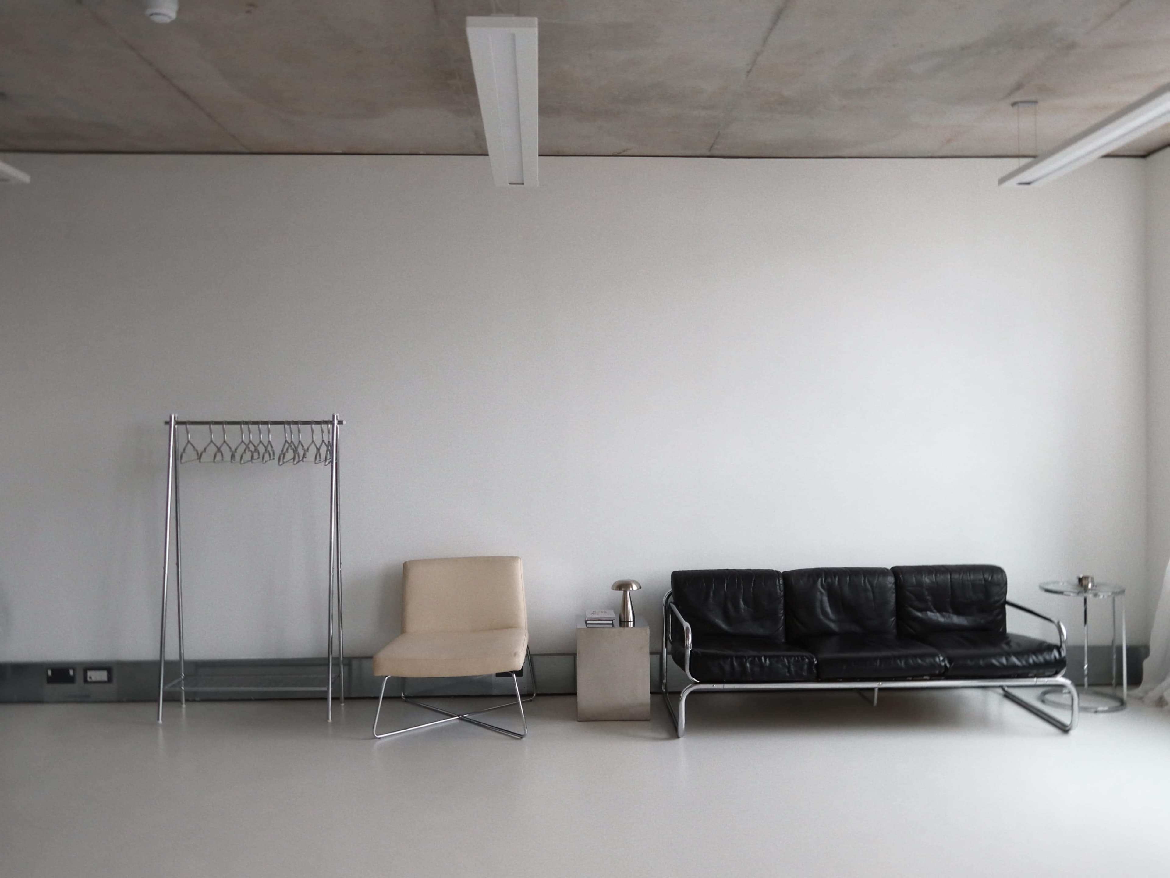 The image shows a minimalist room featuring a black leather sofa, a beige chair, a metal garment rack, and a small side table on a light-colored floor.