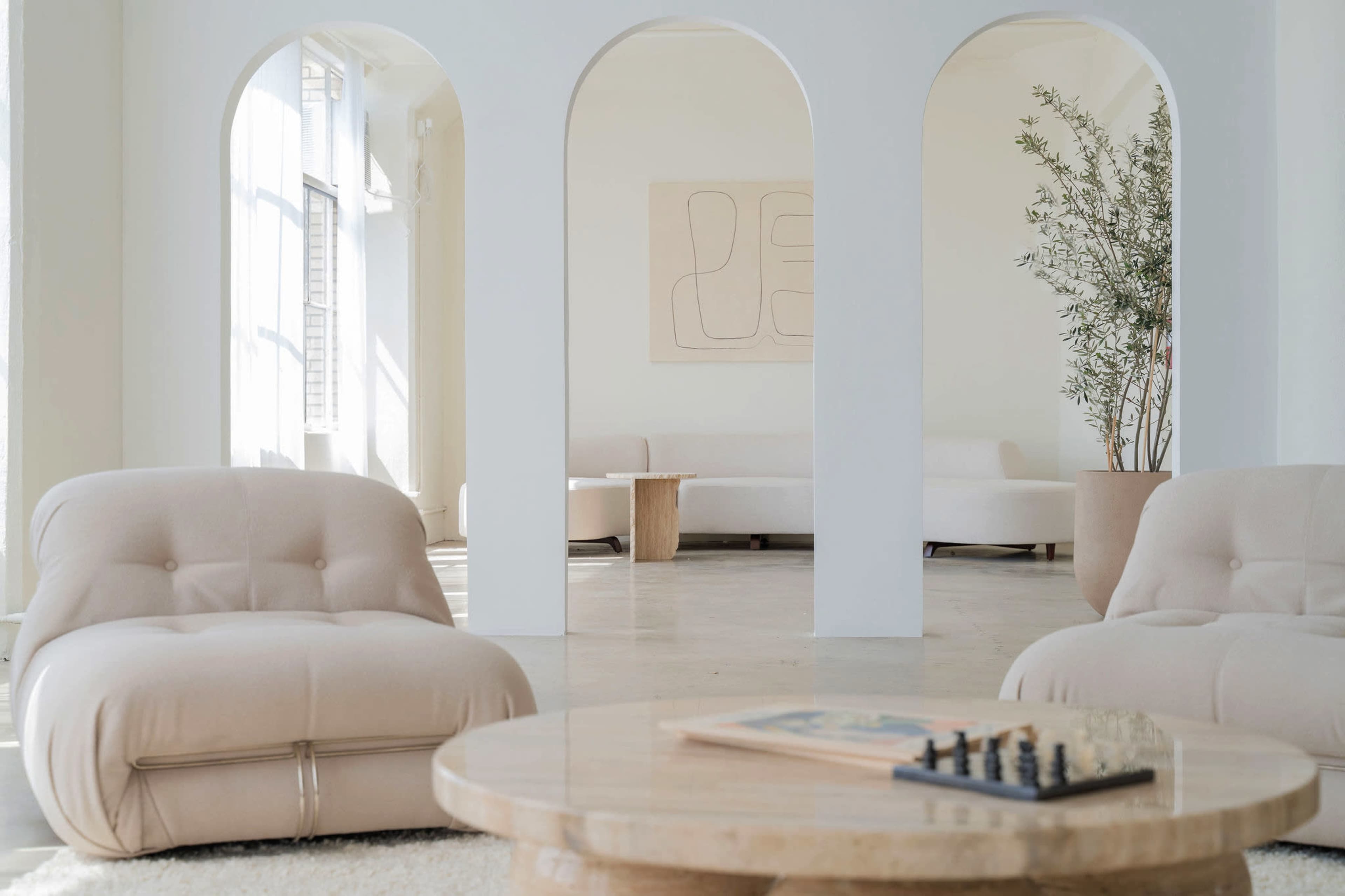 The image shows a spacious, minimalist interior with two plush chairs, a round coffee table, and large archways leading to another area with a sofa and a potted plant.