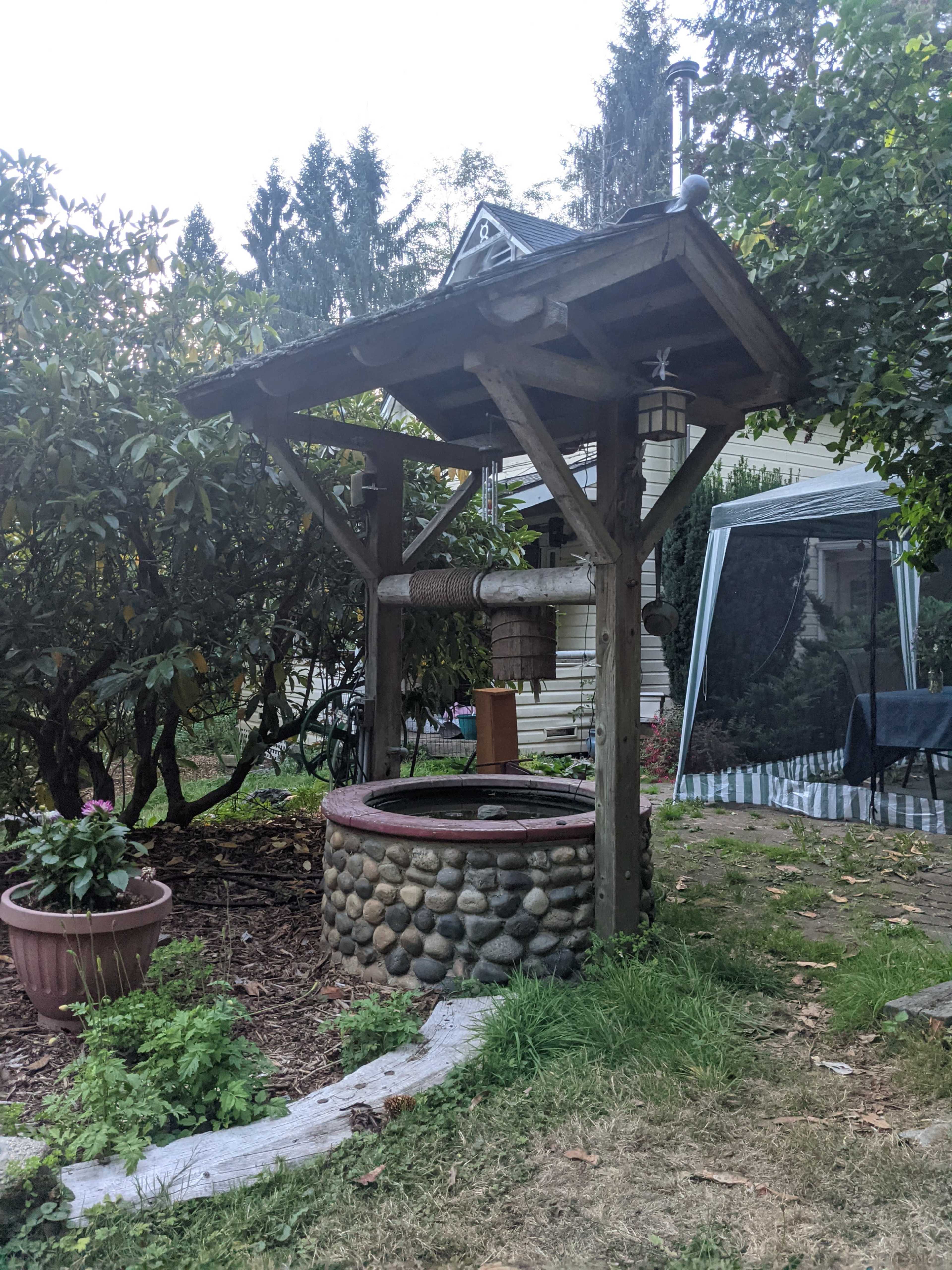 A wooden garden well with a stone base is surrounded by greenery and a fenced area in a residential backyard.