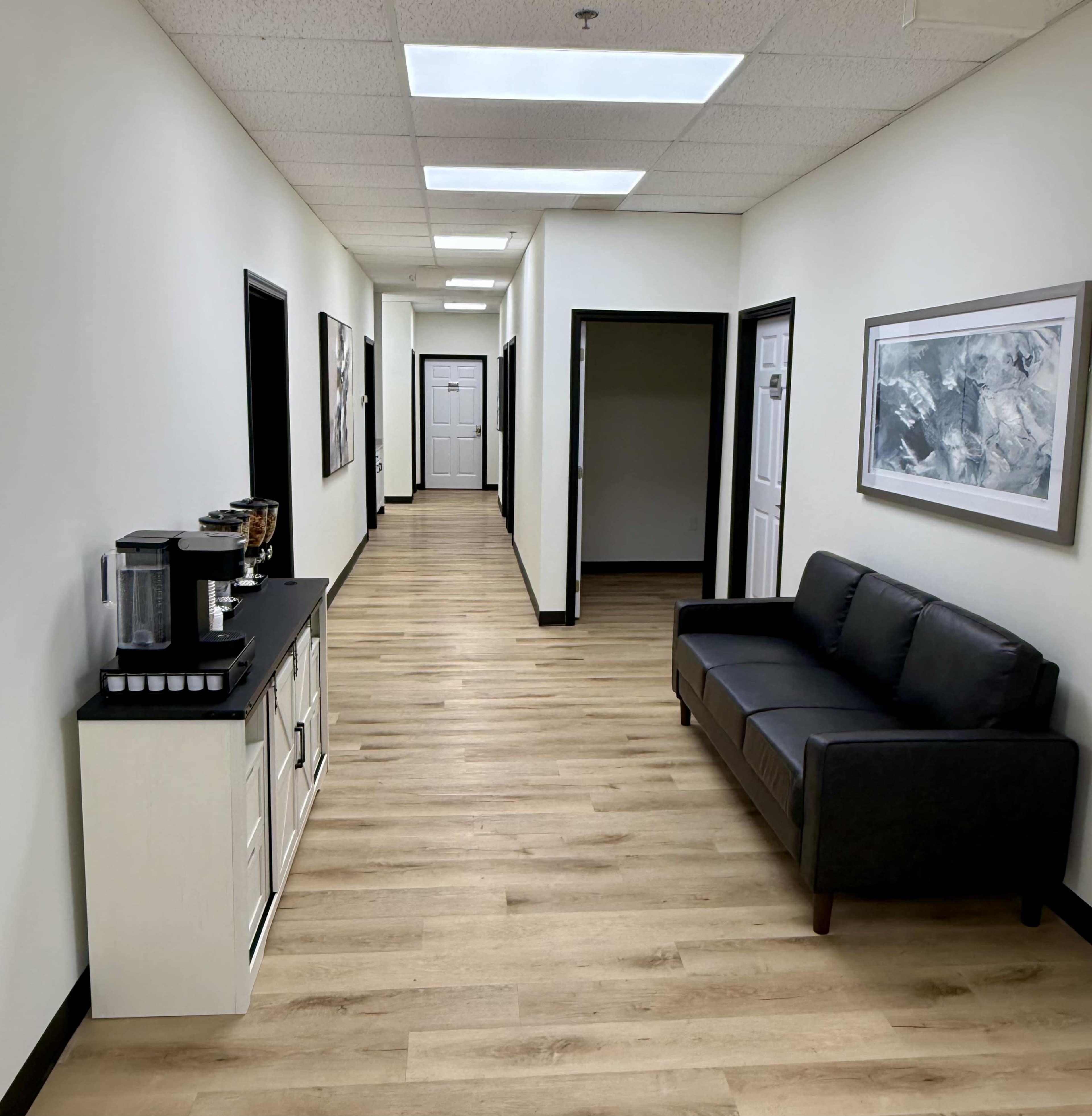 The image shows a well-lit hallway with a dark couch on one side, a coffee station, and several doors along the walls.