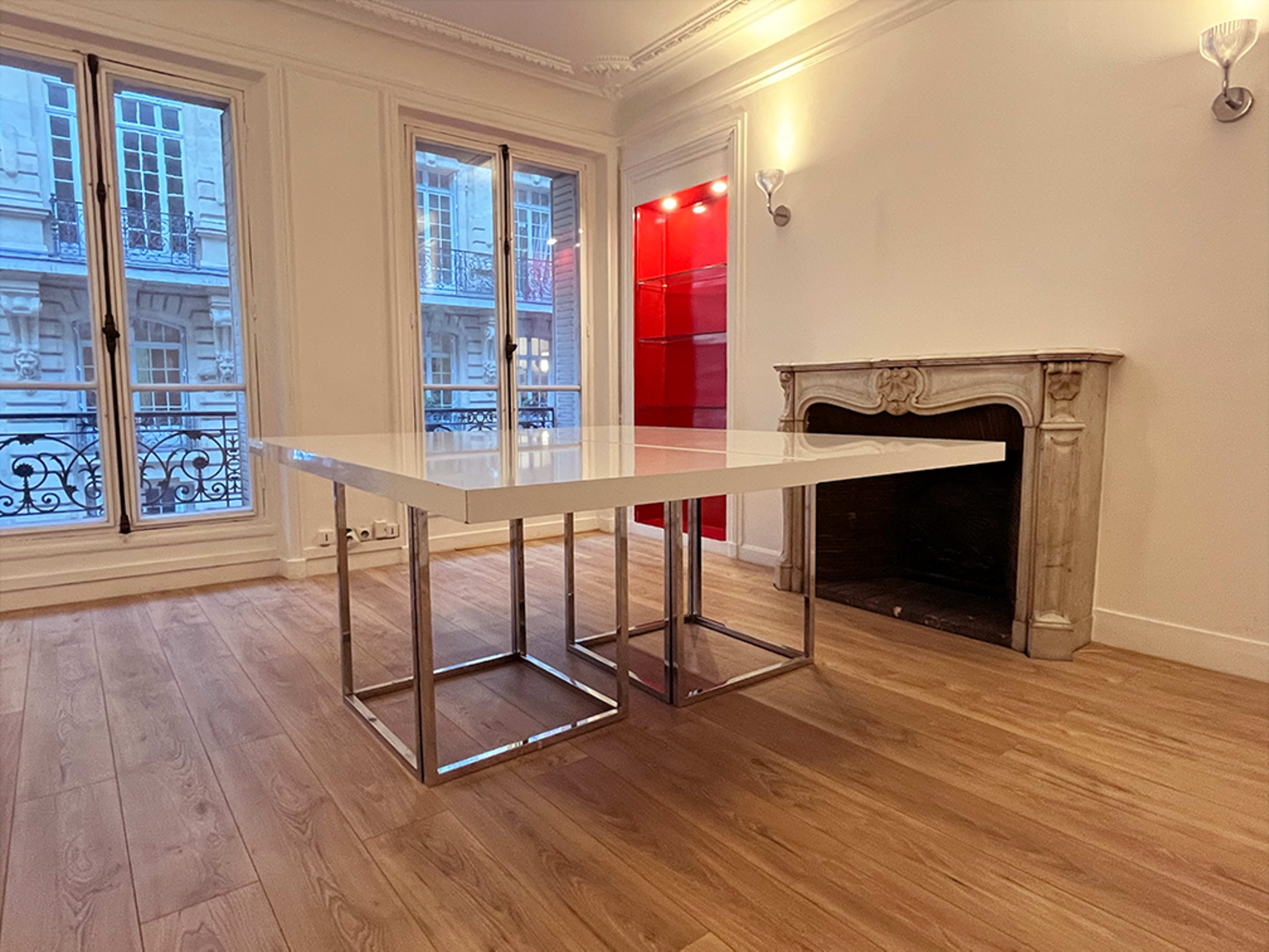 A modern room featuring a white square table with chrome legs, positioned near a fireplace and large windows.