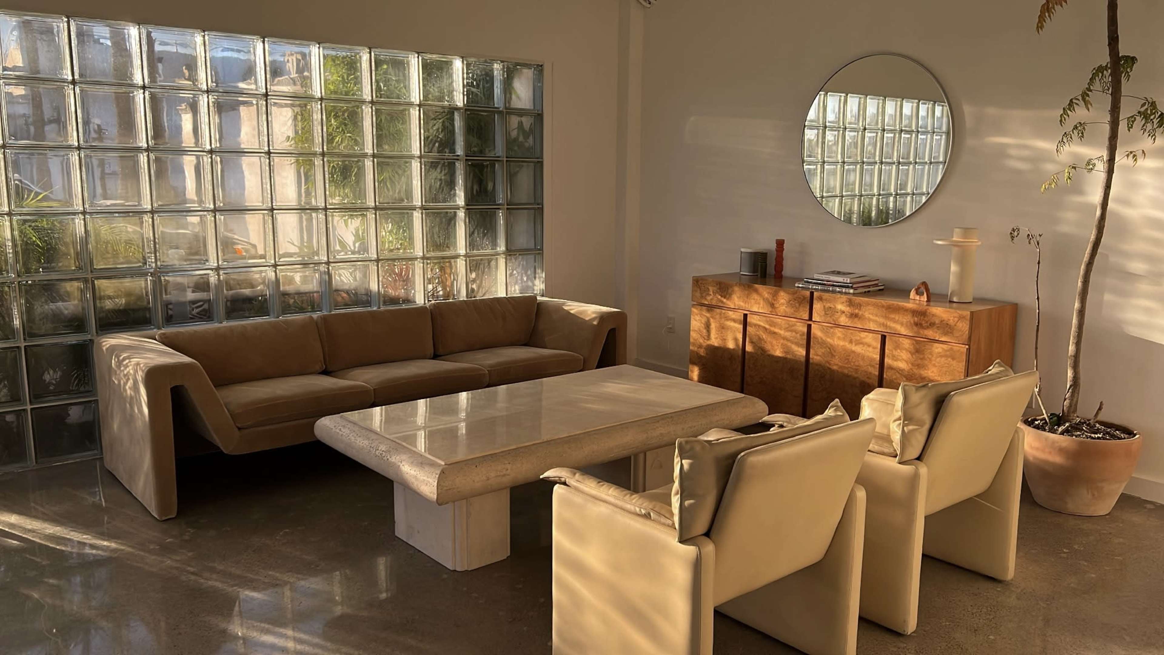 A modern living room with a large beige sofa, a stone coffee table, two light-colored armchairs, and a wooden sideboard, illuminated by natural light from a glass block wall.
