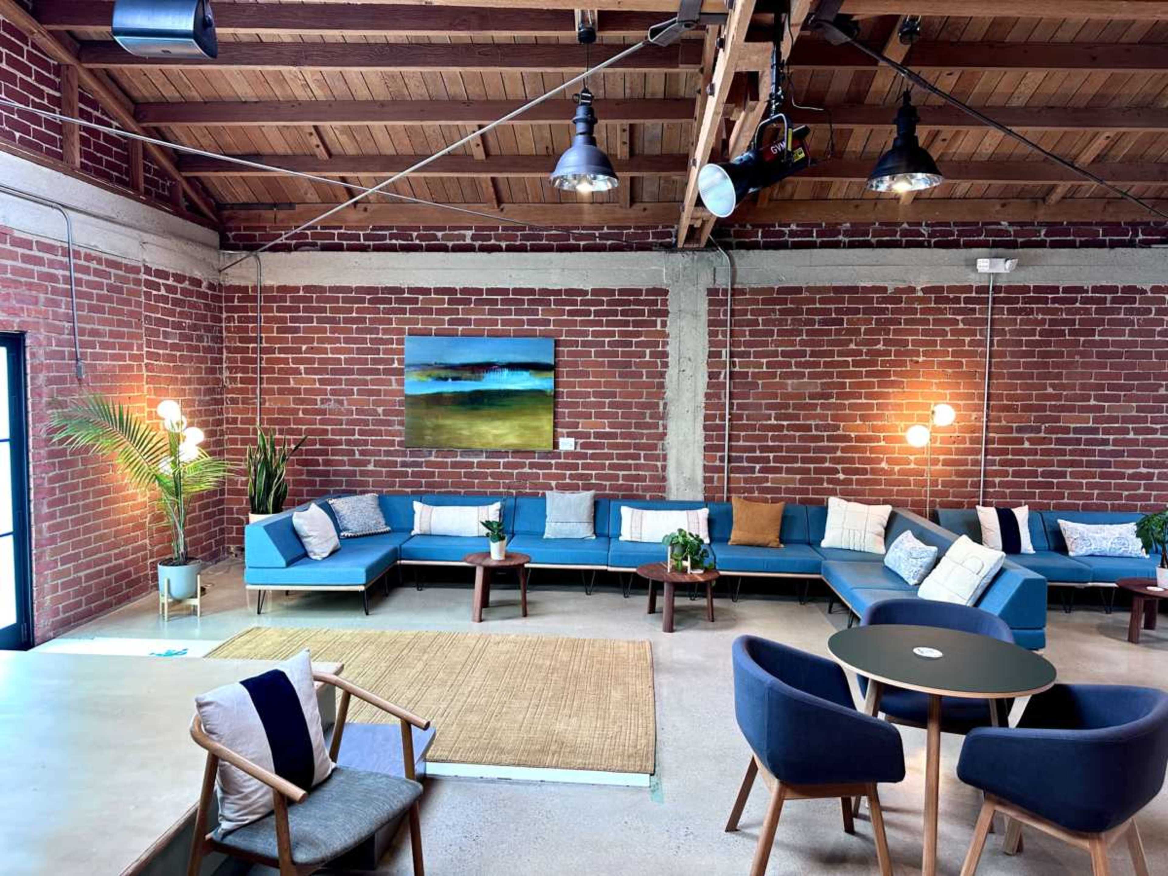 The image shows a spacious interior with a mix of blue couches, a round table, and stone flooring, framed by exposed brick walls and wooden beams.