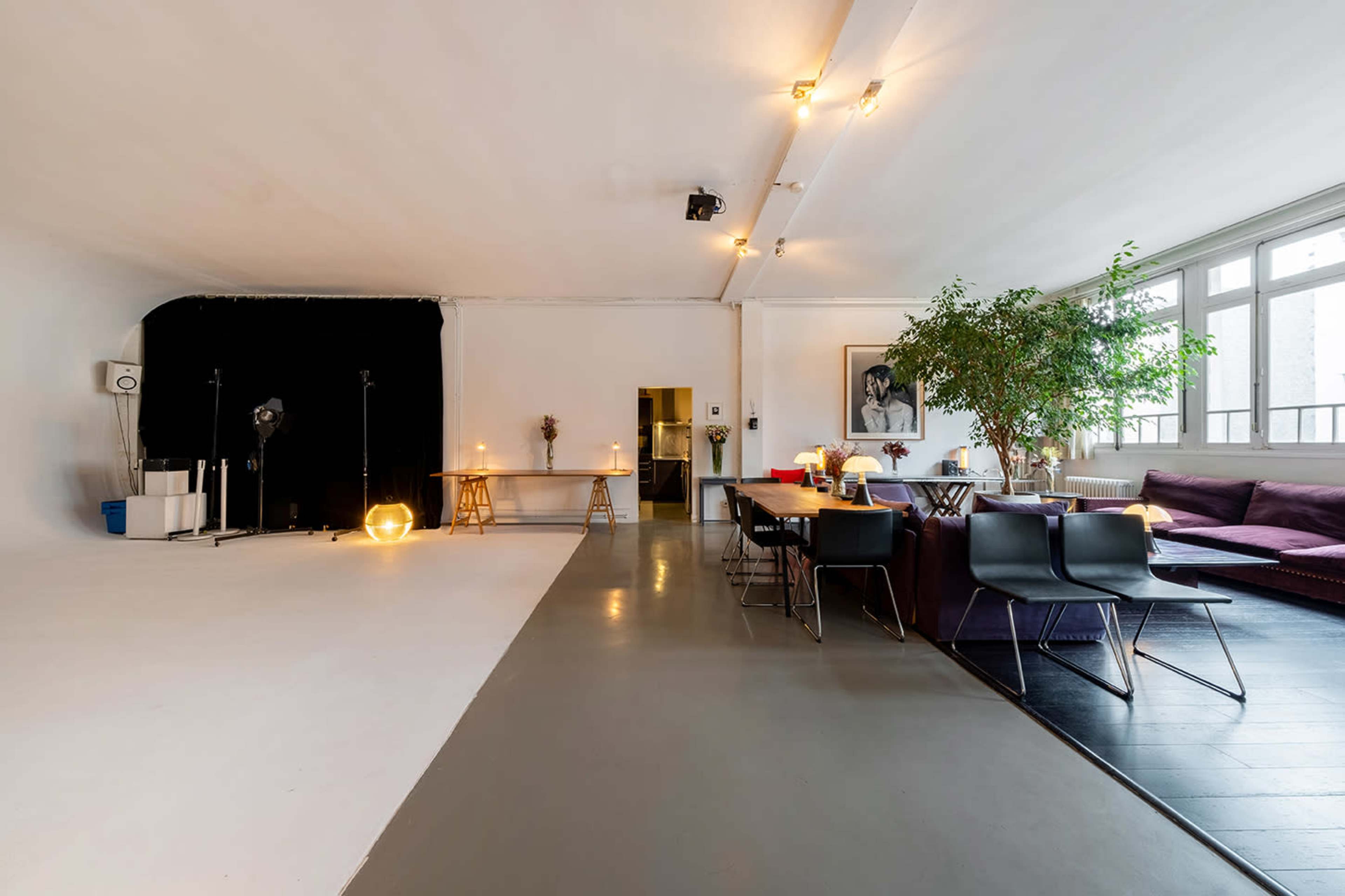 A spacious studio with a black backdrop, a dining area with chairs, a table, and a couch, illuminated by overhead lights.