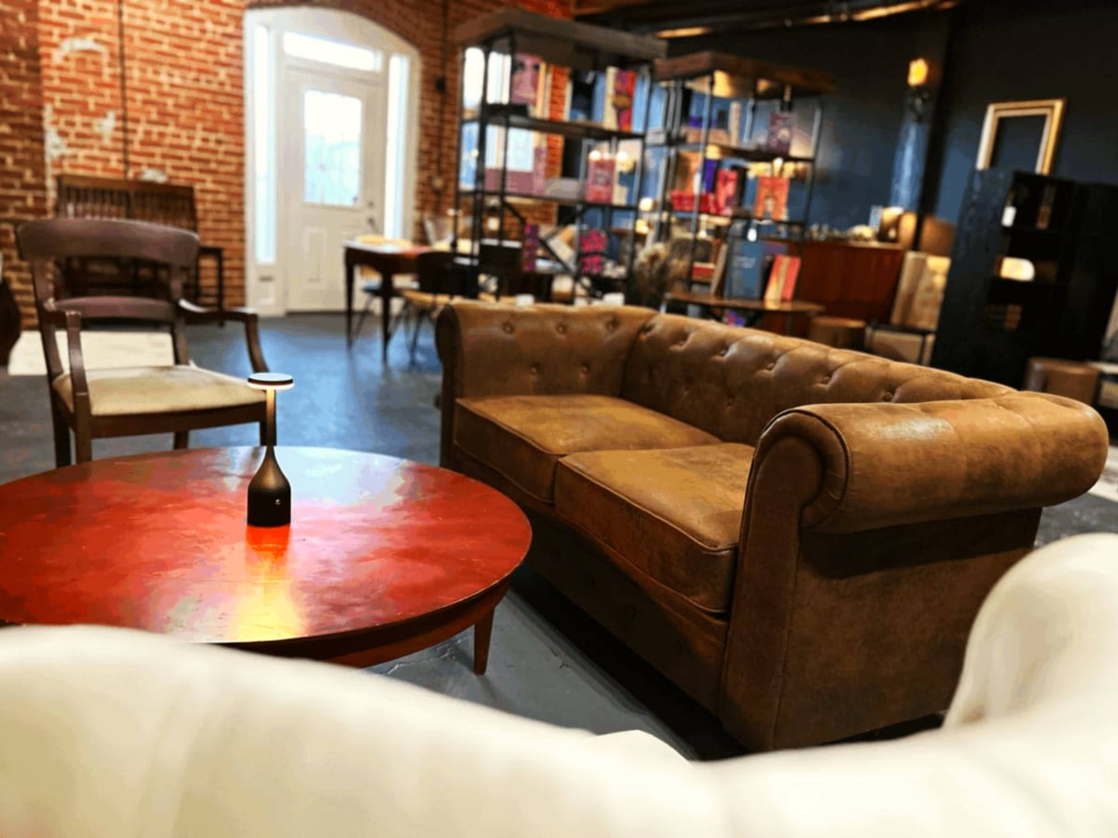 The image shows a cozy seating area featuring a brown leather chesterfield sofa, a round red coffee table with a vase, and wooden chairs against a backdrop of exposed brick walls and bookshelves.