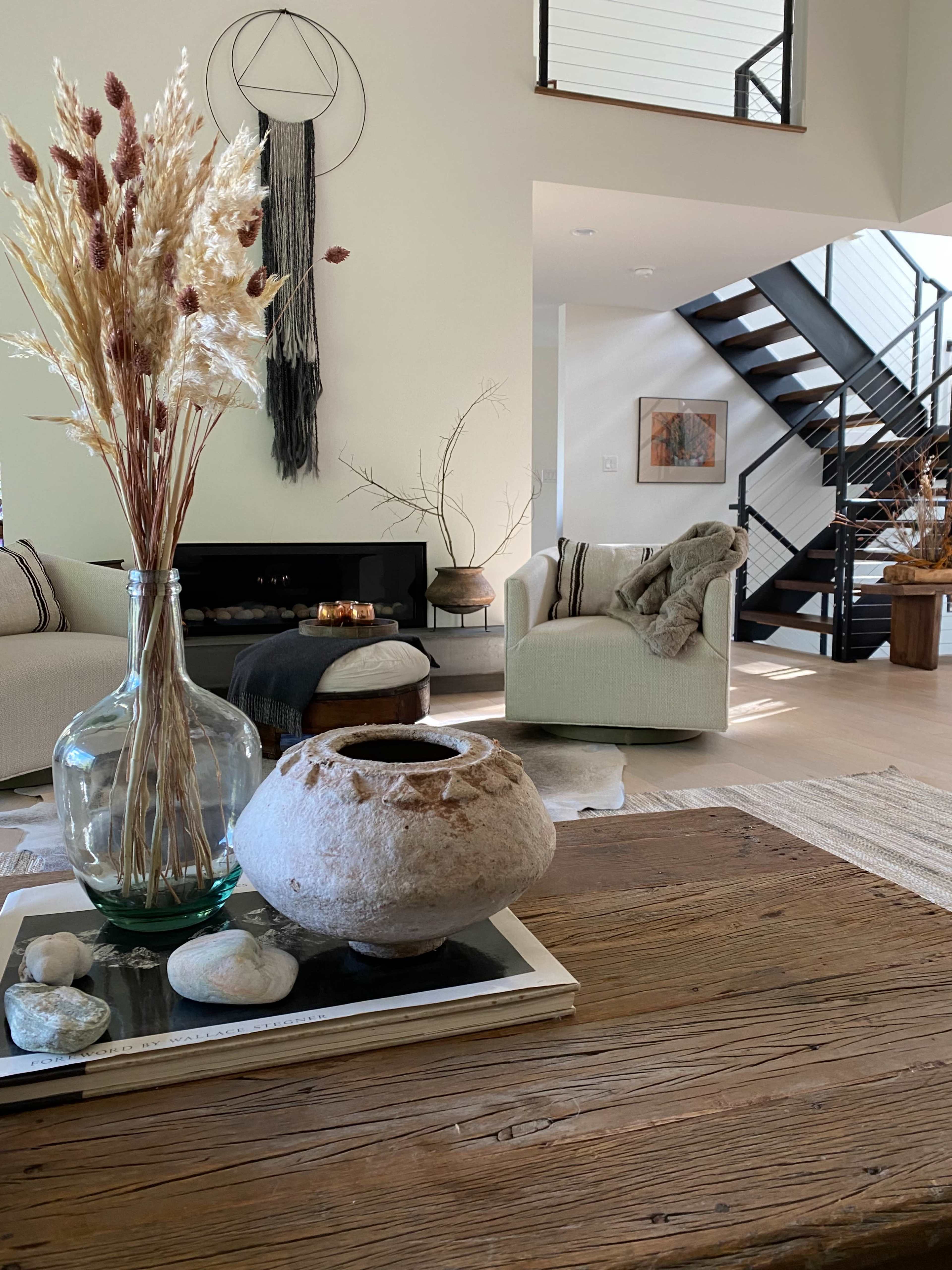 A modern living room with a wooden coffee table featuring a decorative bowl, a vase of dried flowers, and a staircase in the background.